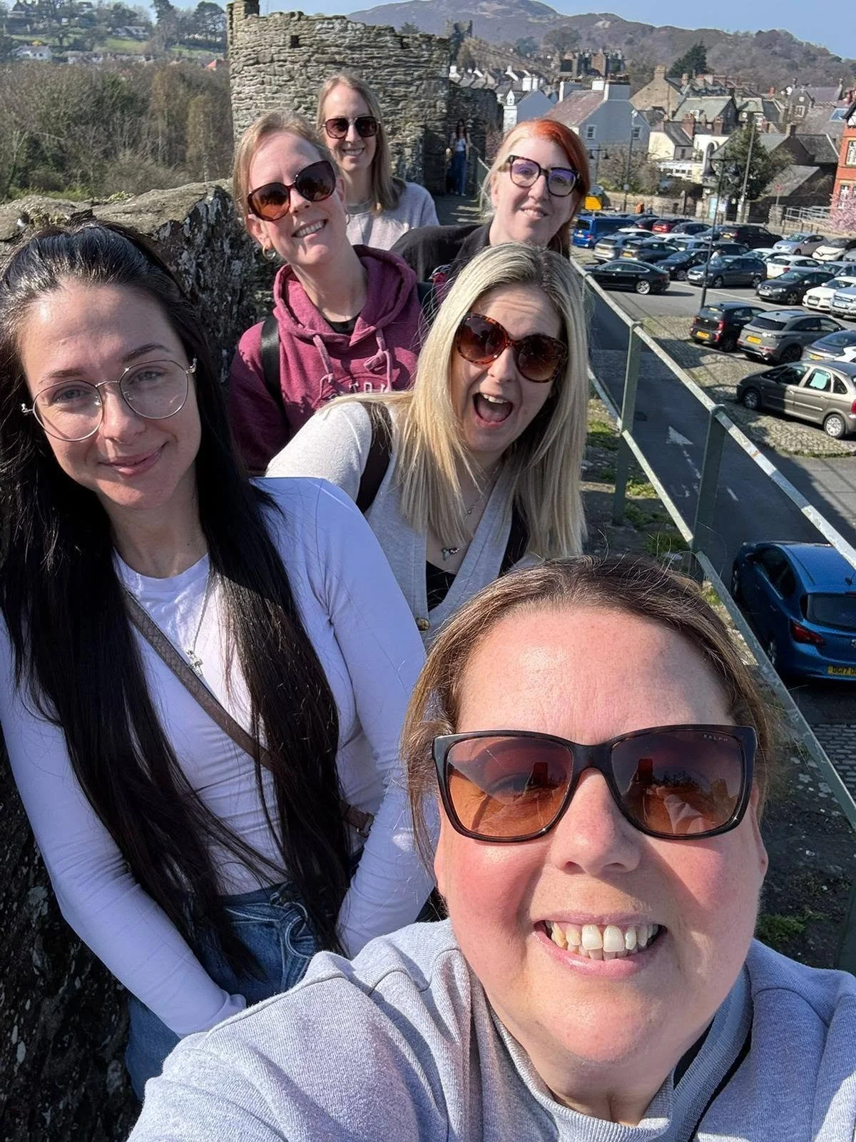 Group of six women taking a selfie on a balcony with historic stone walls, a parking lot, and a town with houses and mountains in the background.