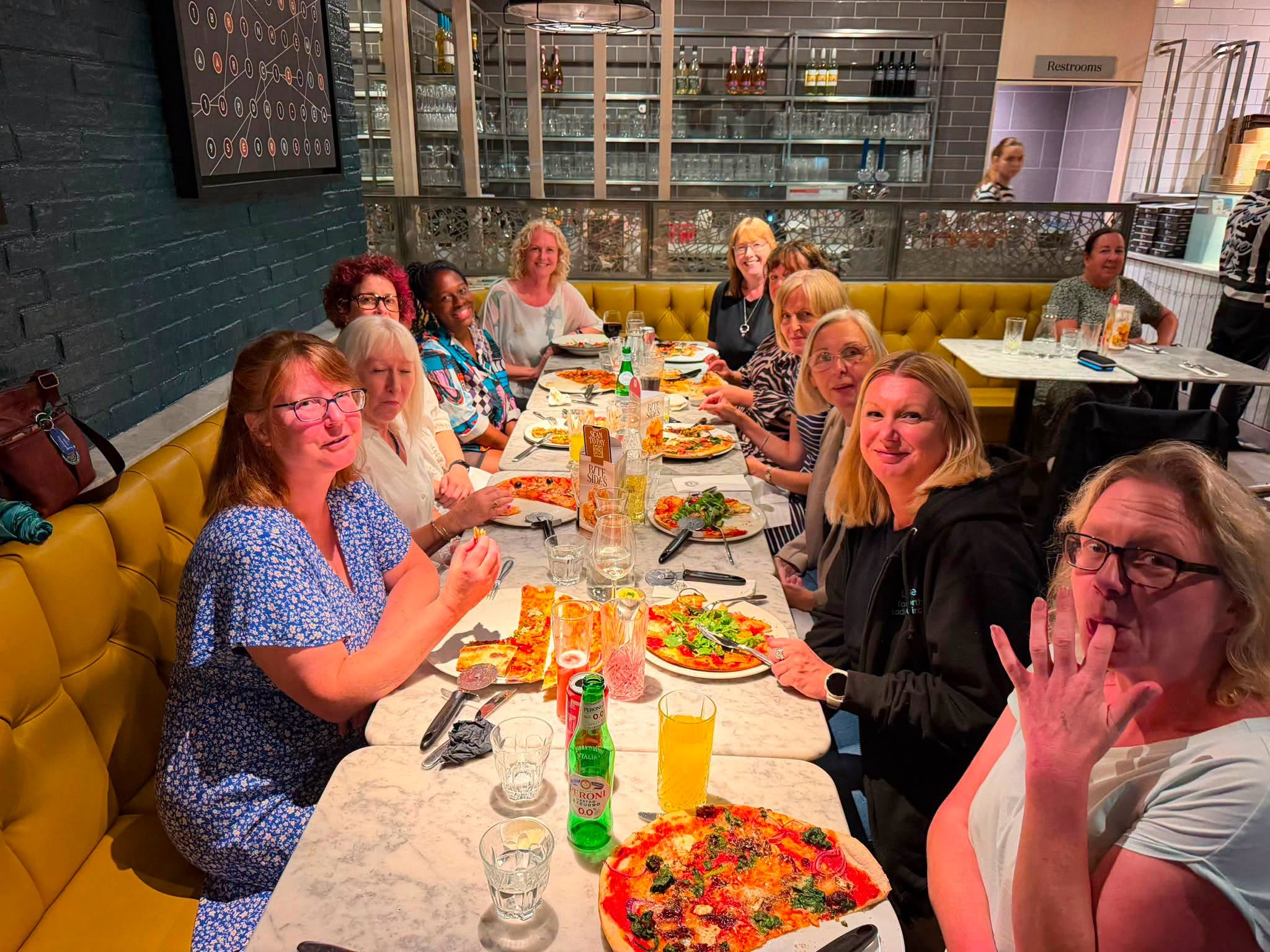A group of women dining together at a restaurant table, with pizza, drinks, and cutlery, seated against a yellow booth with a dark brick wall and shelves of glassware behind them.
