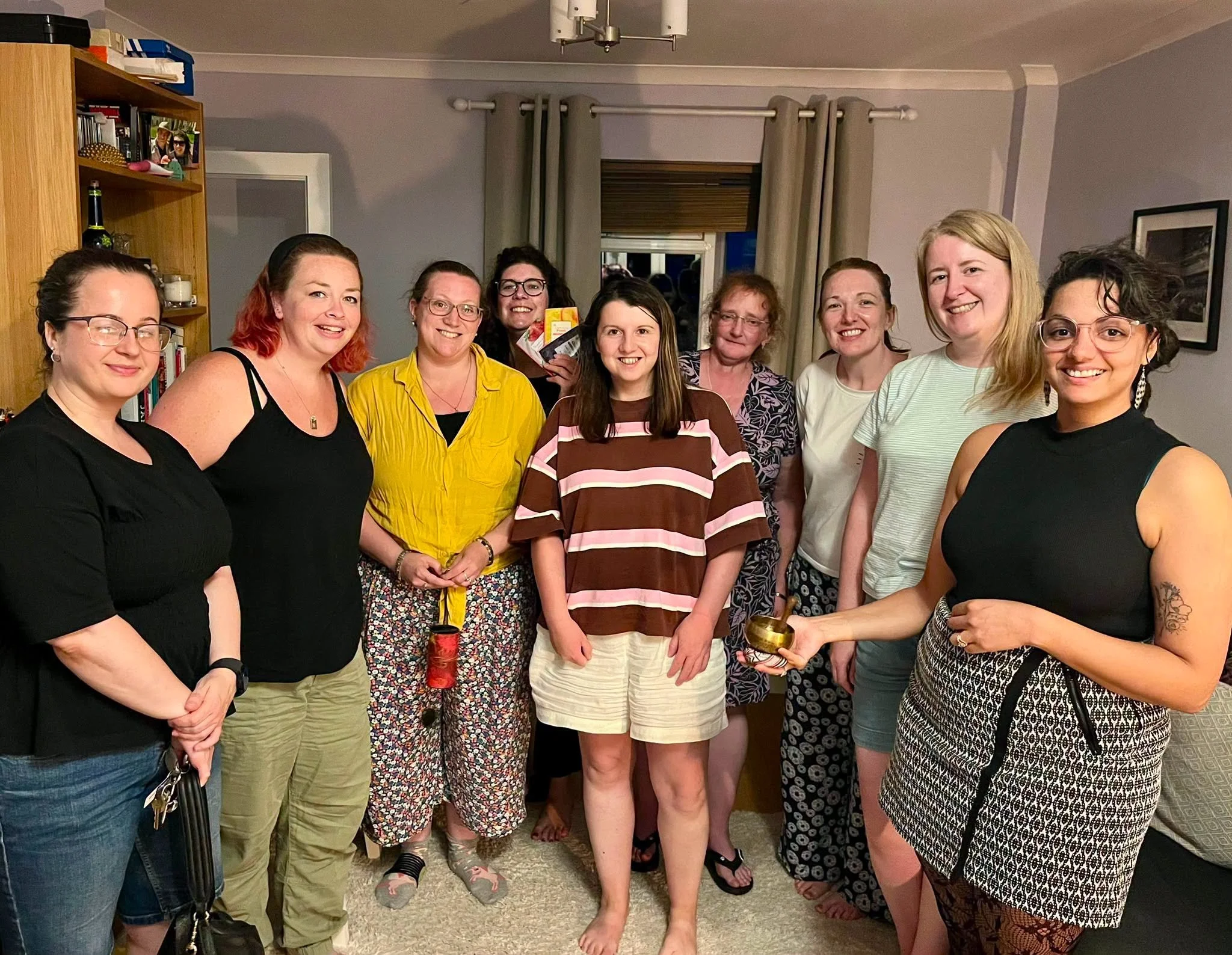 Ten women are gathered in a living room, smiling at the camera. They are standing close together, some with glasses and wearing casual clothing. The background includes a bookshelf with books and decorative items, a window with curtains, and a framed picture on the wall.