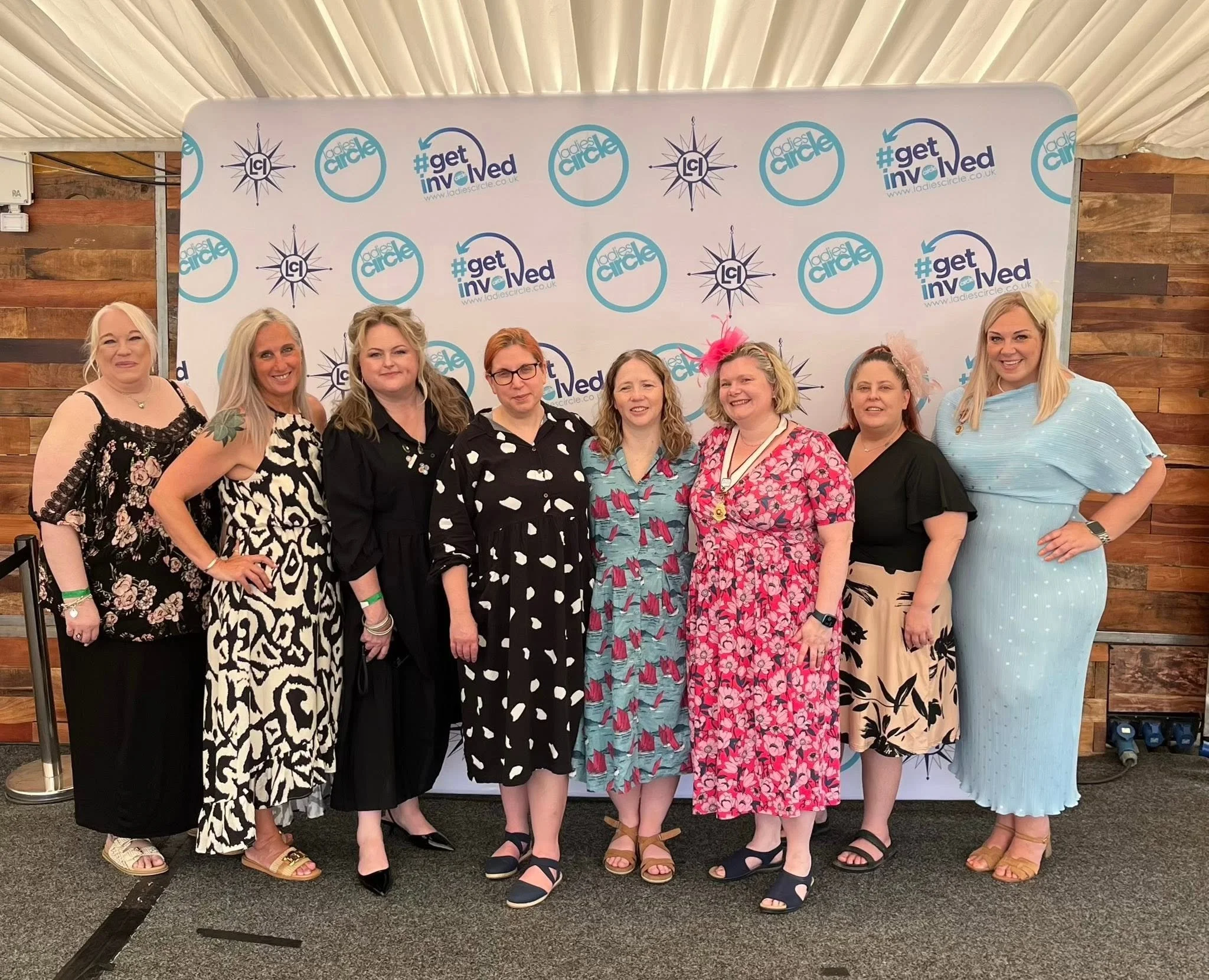 Group of women standing in front of a backdrop with logos that say "Ladies Circle" and "#getinvolved" at an event. They are wearing colorful dresses and smiling.
