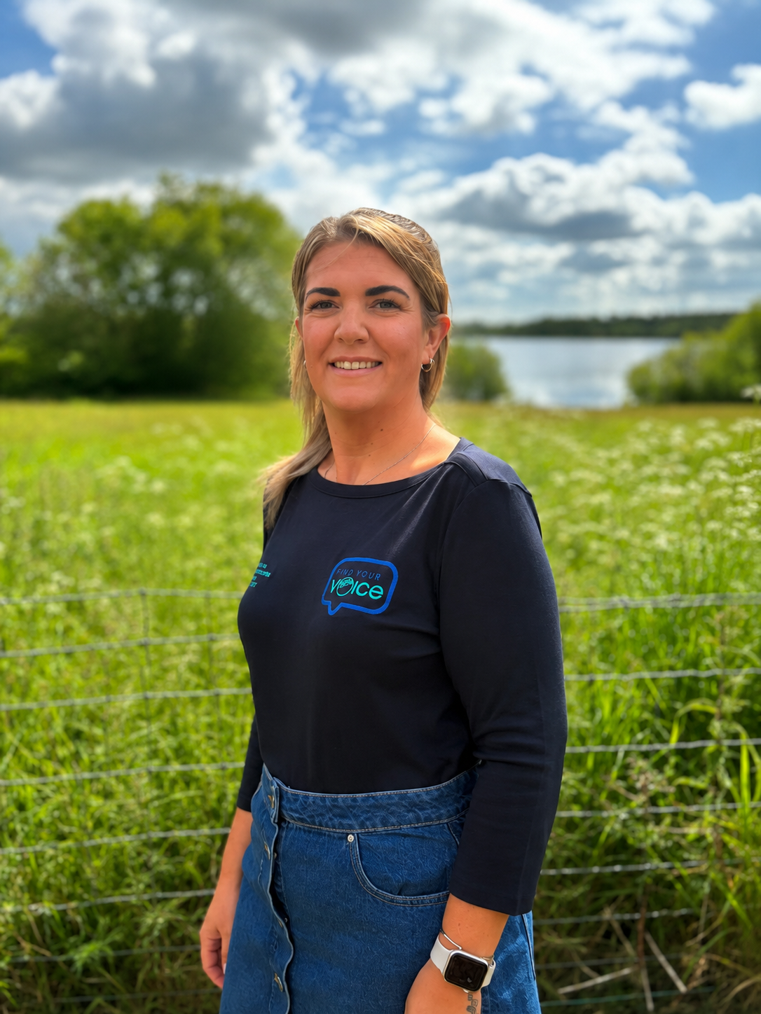 A woman standing outdoors in a field with green grass and blooming wildflowers, smiling, wearing a black t-shirt with a speech bubble logo, blue jeans, and a smartwatch, with a lake, trees, and a partly cloudy sky in the background.