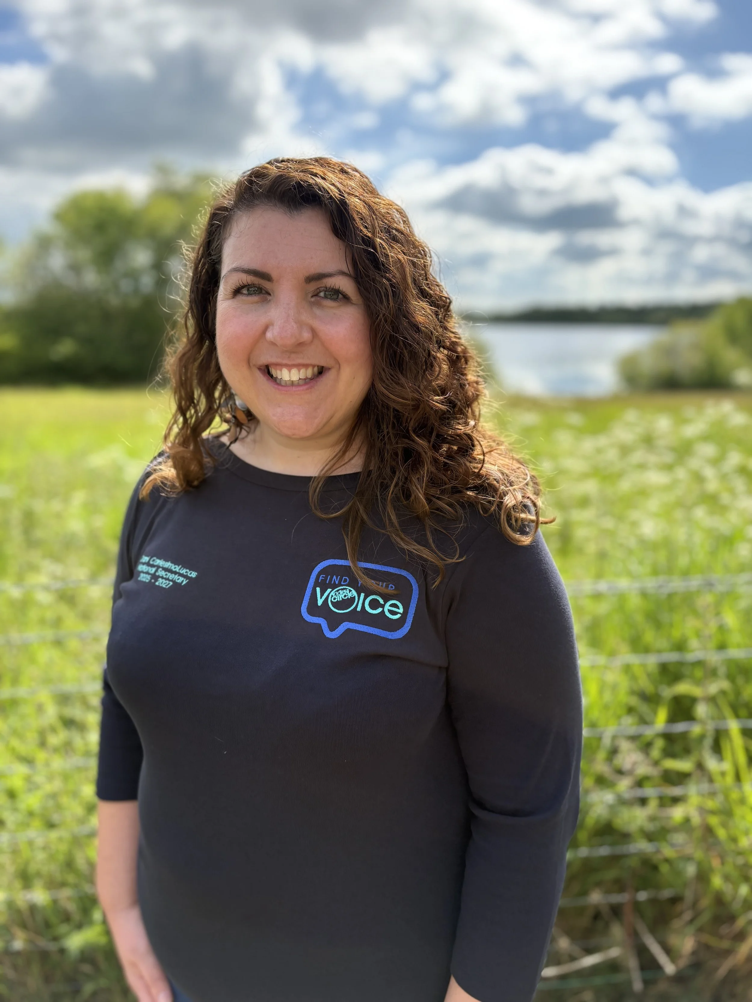 A woman with curly brown hair and light skin smiling outdoors with a river, green trees, and a partly cloudy sky in the background.