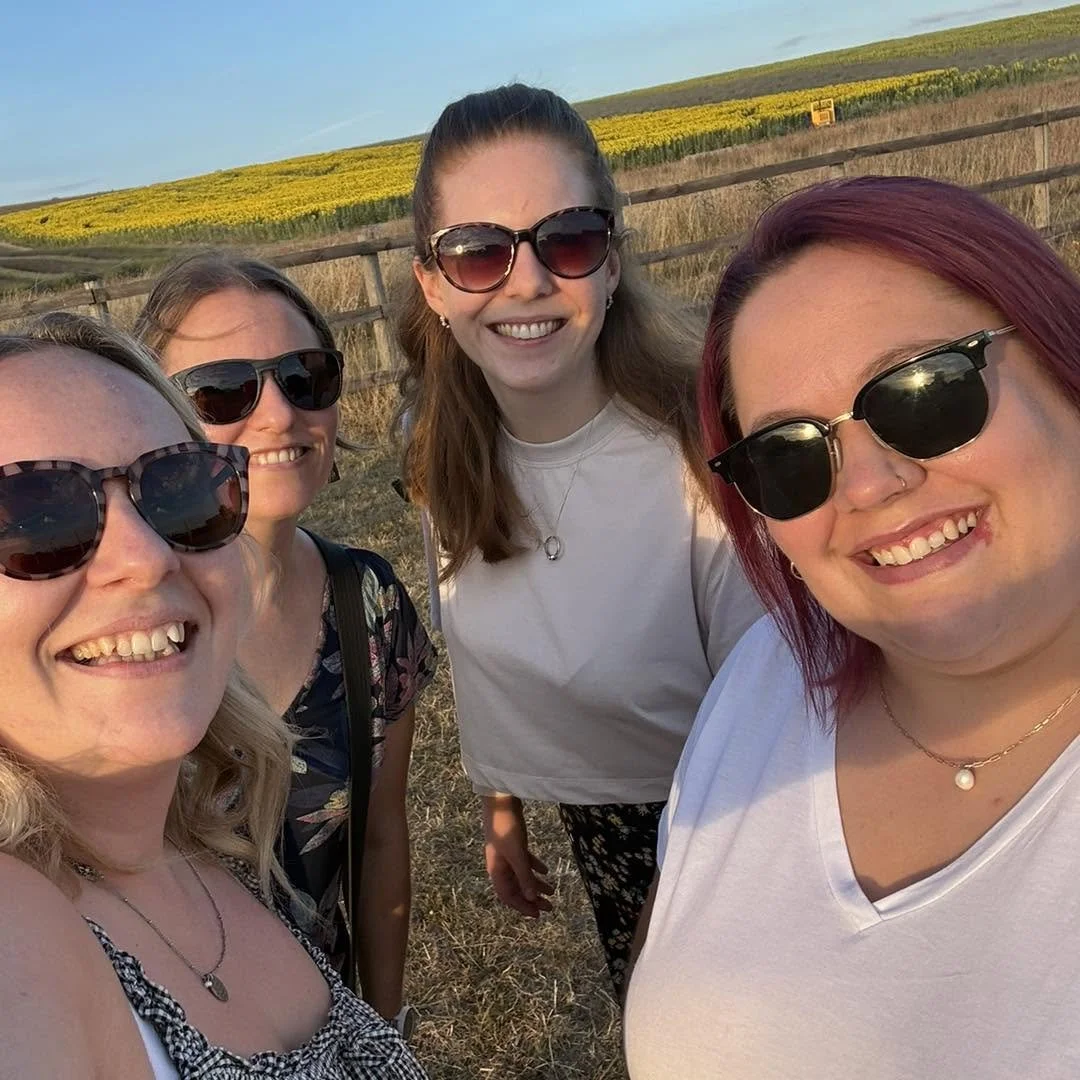 Four women smiling and taking a selfie outdoors at sunset, wearing sunglasses, with a fence, fields, and a hill with yellow flowers in the background.