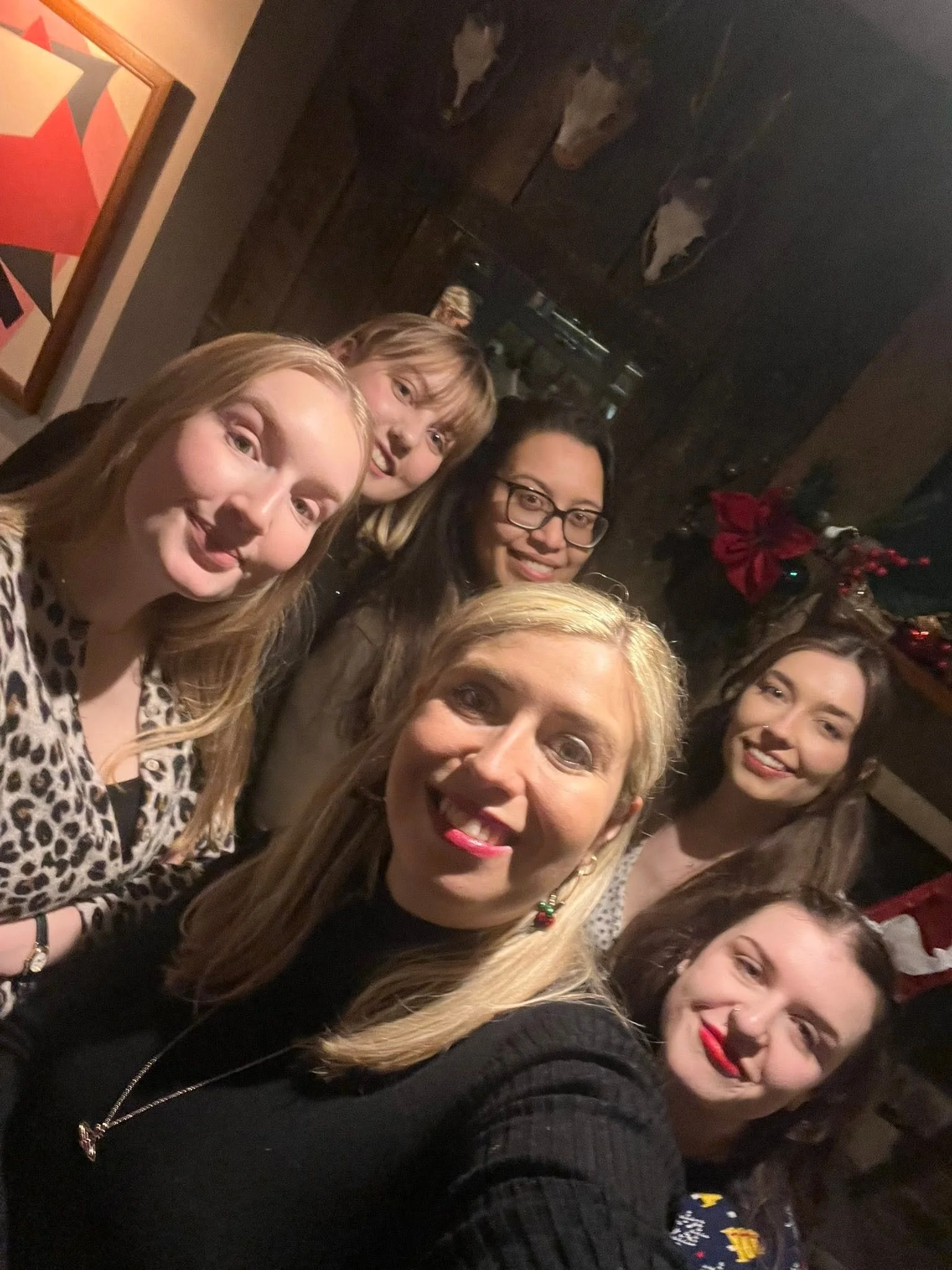 A group of women smiling and taking a selfie in a dimly lit room decorated with Christmas ornaments and poinsettias.