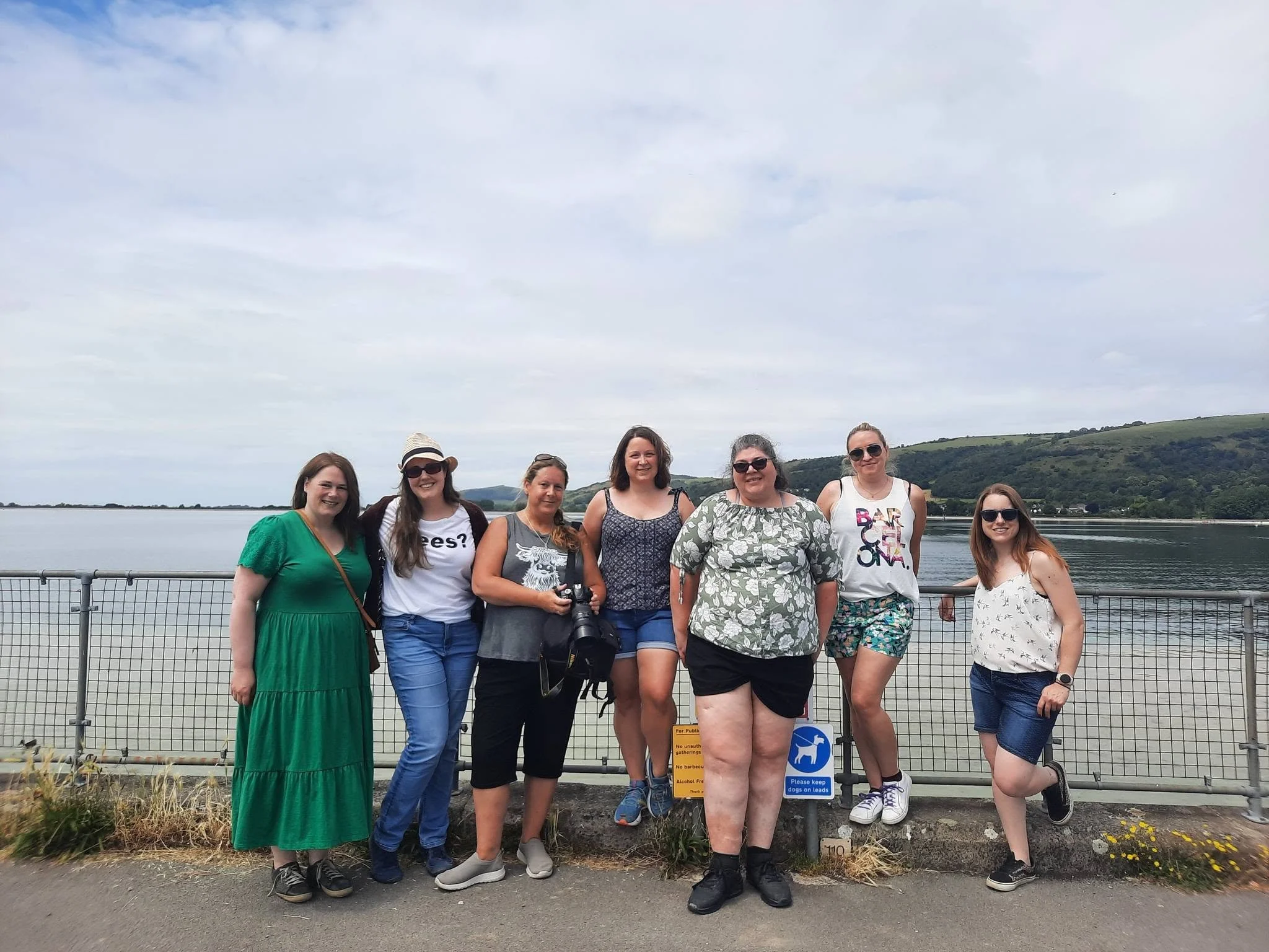A group of nine women standing on a waterfront promenade with hills and water in the background, some wearing sunglasses and casual summer clothing, and a sign that says 'Please keep dogs on leads' in the foreground.