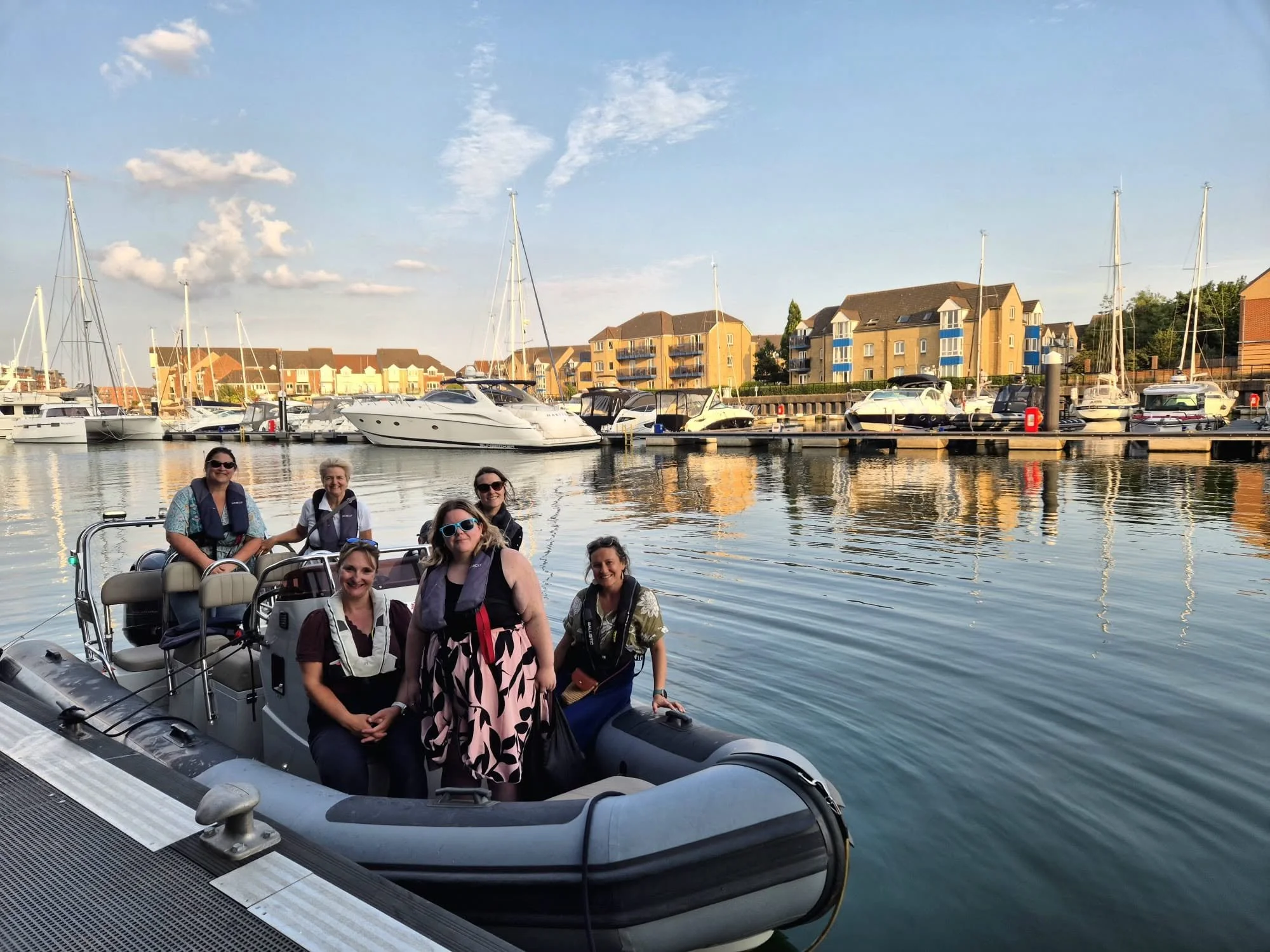 A group of people on a boat in a marina with sailboats and yachts, residential buildings, and a blue sky with clouds in the background.
