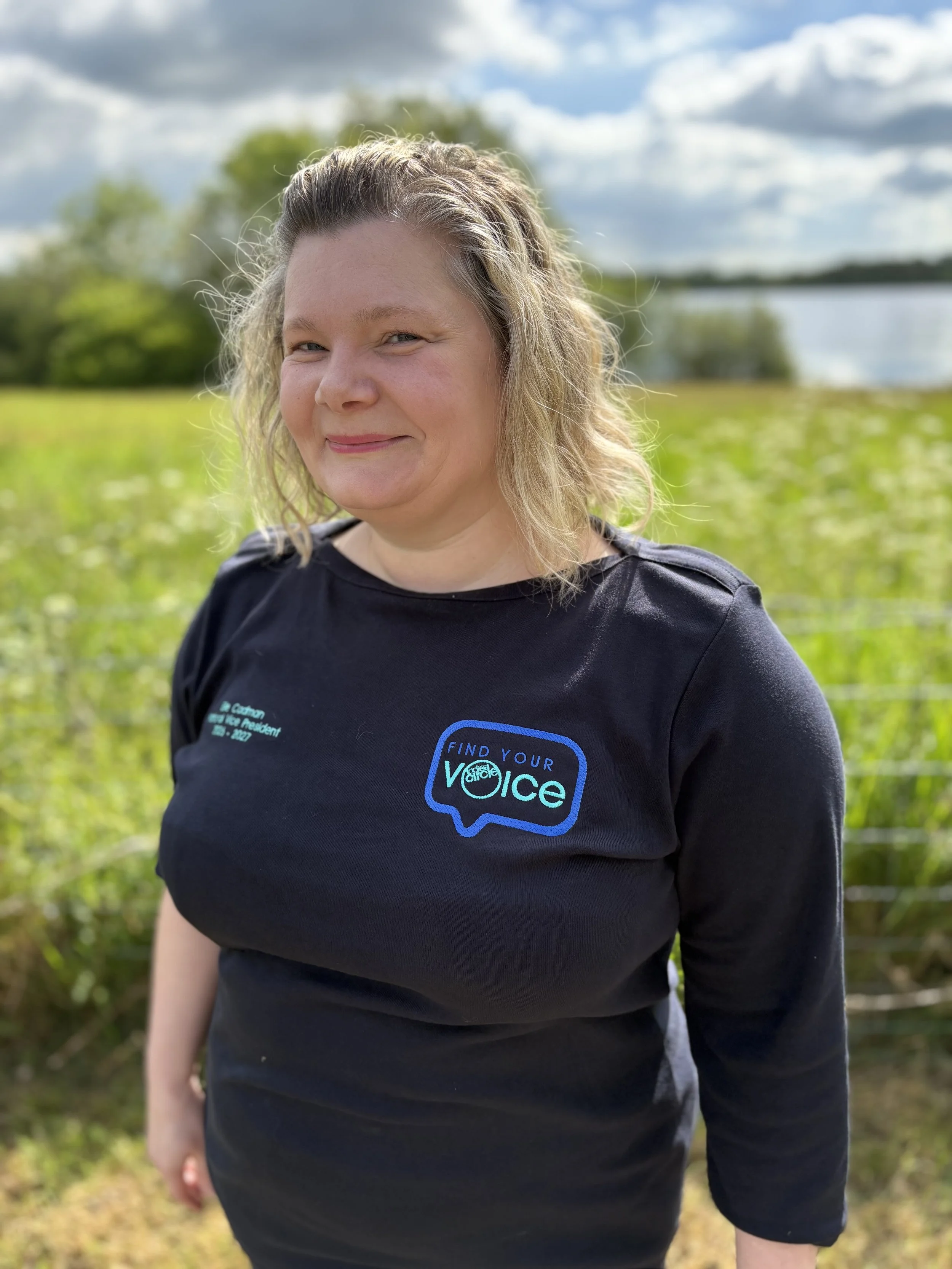 A woman with blonde hair and light skin smiling outdoors, wearing a black t-shirt with a logo that says 'Find Your Voice' and a speech bubble.