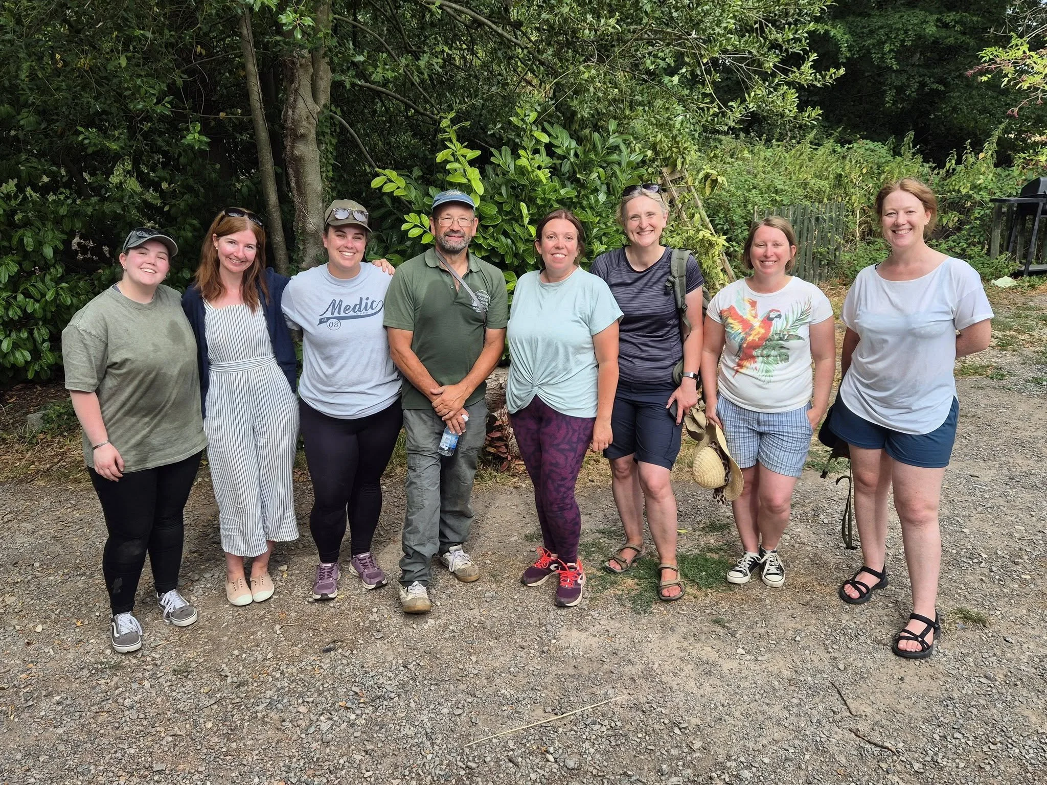 A group of eight smiling people standing outdoors on a dirt path in front of green bushes, wearing casual clothing and sneakers, with some carrying backpacks and sunglasses.