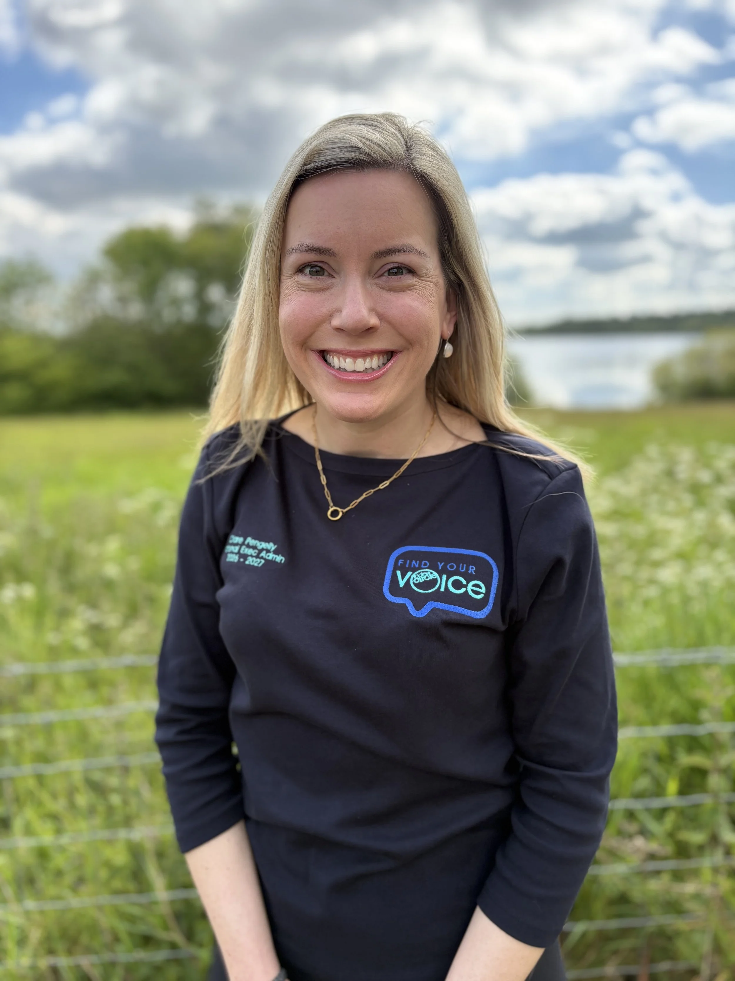 A smiling woman with blonde hair wearing black clothing and a gold necklace, standing outdoors with greenery and a body of water in the background under a partly cloudy sky.