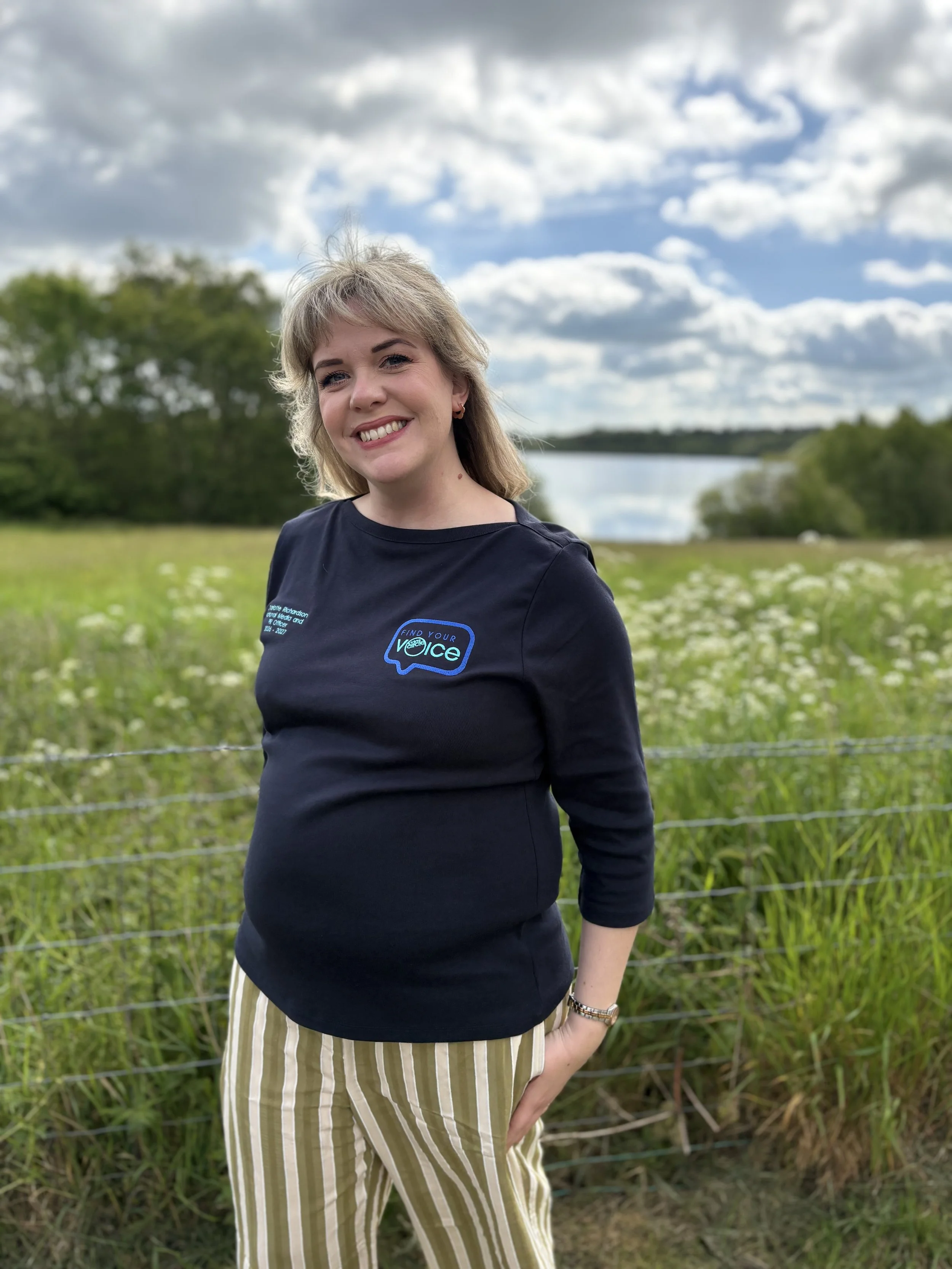 A smiling woman standing outdoors near a fence in a grassy field with trees and a river in the background under a cloudy sky.