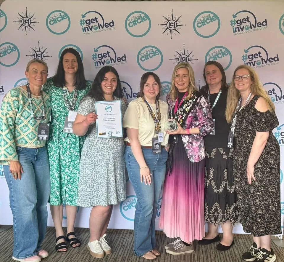 Group of eight women at an event, with a backdrop displaying logos and hashtags for 'Ladies Circle' and '#getinvolved.' One woman is holding a plaque, and another is holding an award.