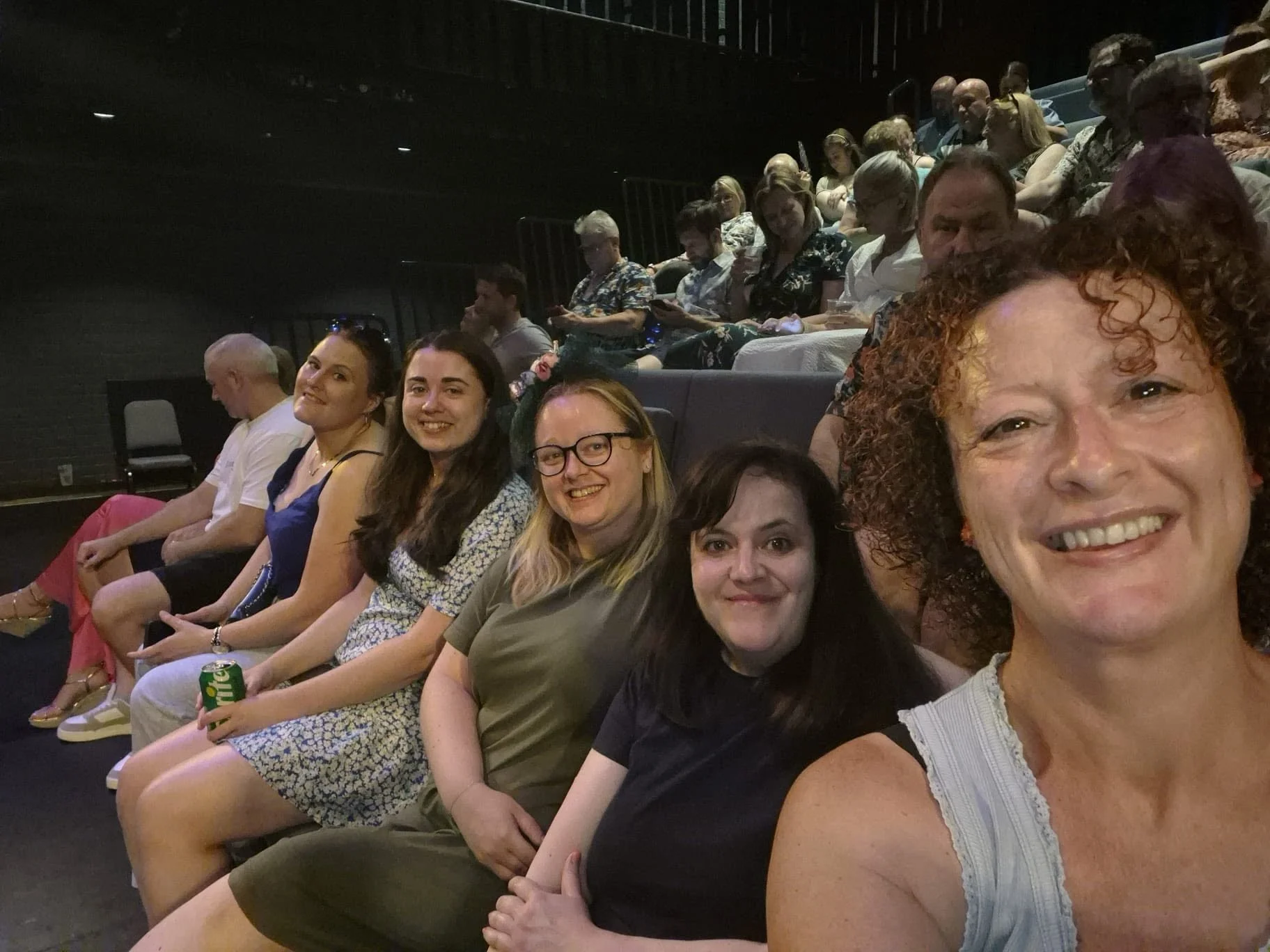 A group of smiling women and a few men sitting in theater seats, taking a selfie, with more people seated in the background.
