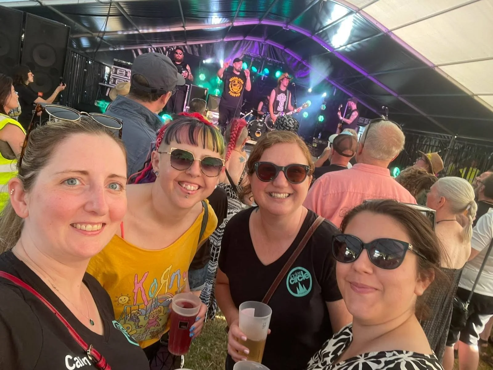 A group of four smiling women wearing sunglasses at a music festival or concert, with a stage and band performing in the background, under a large tent with colorful lights.