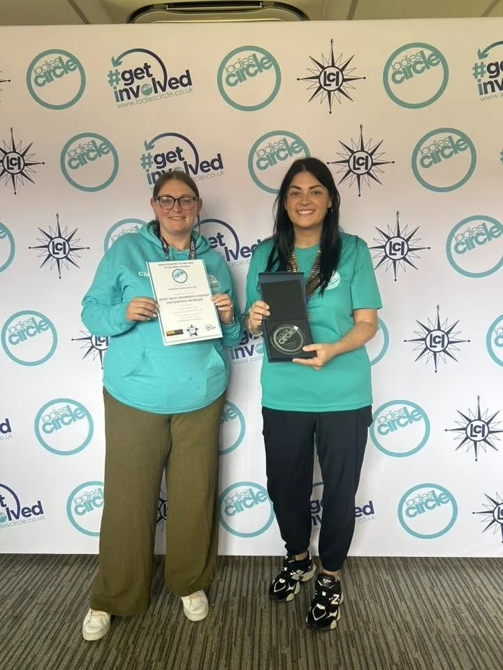 Two women standing in front of a decorated backdrop with logos and hashtags. They are smiling, holding a certificate and a box with a medal or award. Both are wearing teal shirts, suggesting they are part of an event or organization, possibly related to LADIES CIRCLE.