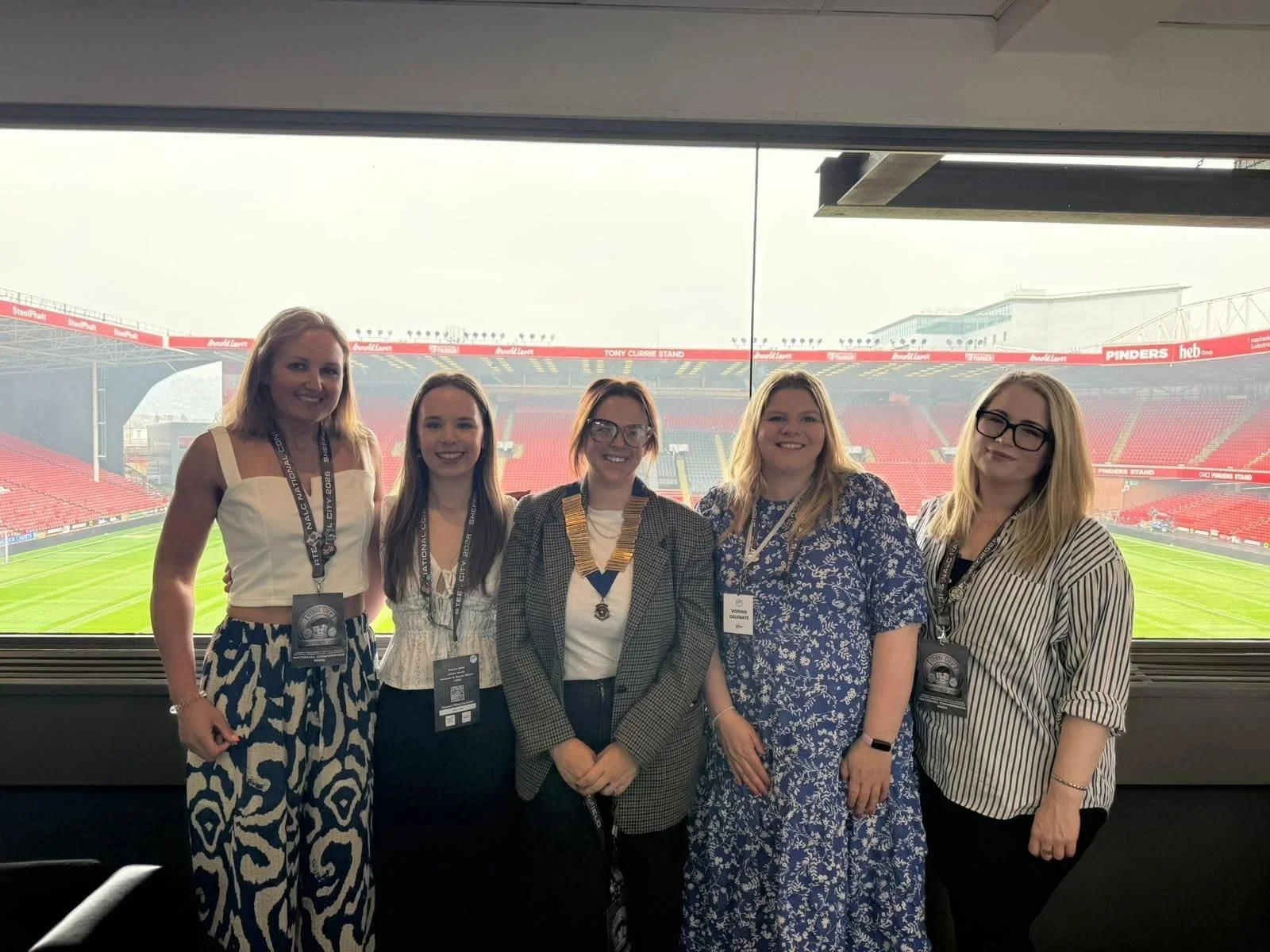 Five women standing inside a sports arena, with green football field visible through large window behind them.
