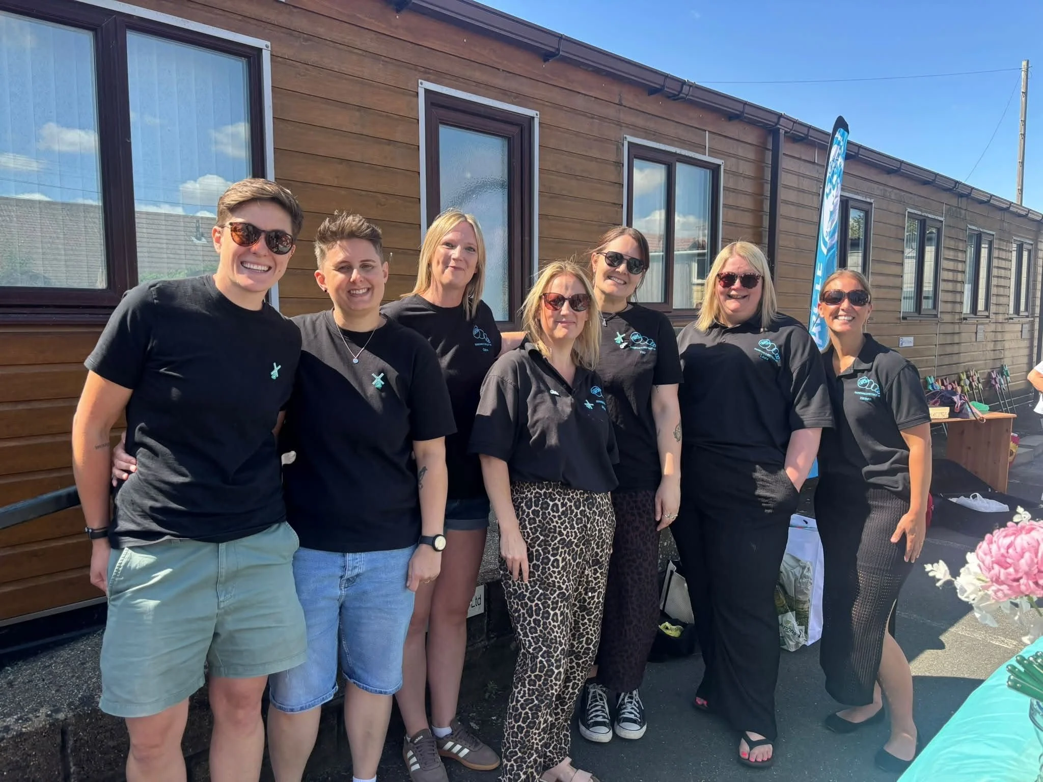 A group of seven women standing in front of a brown wooden building. They are smiling and wearing black shirts, some with logos, and sunglasses. There are tables with pink flowers and a blue flag to the side.