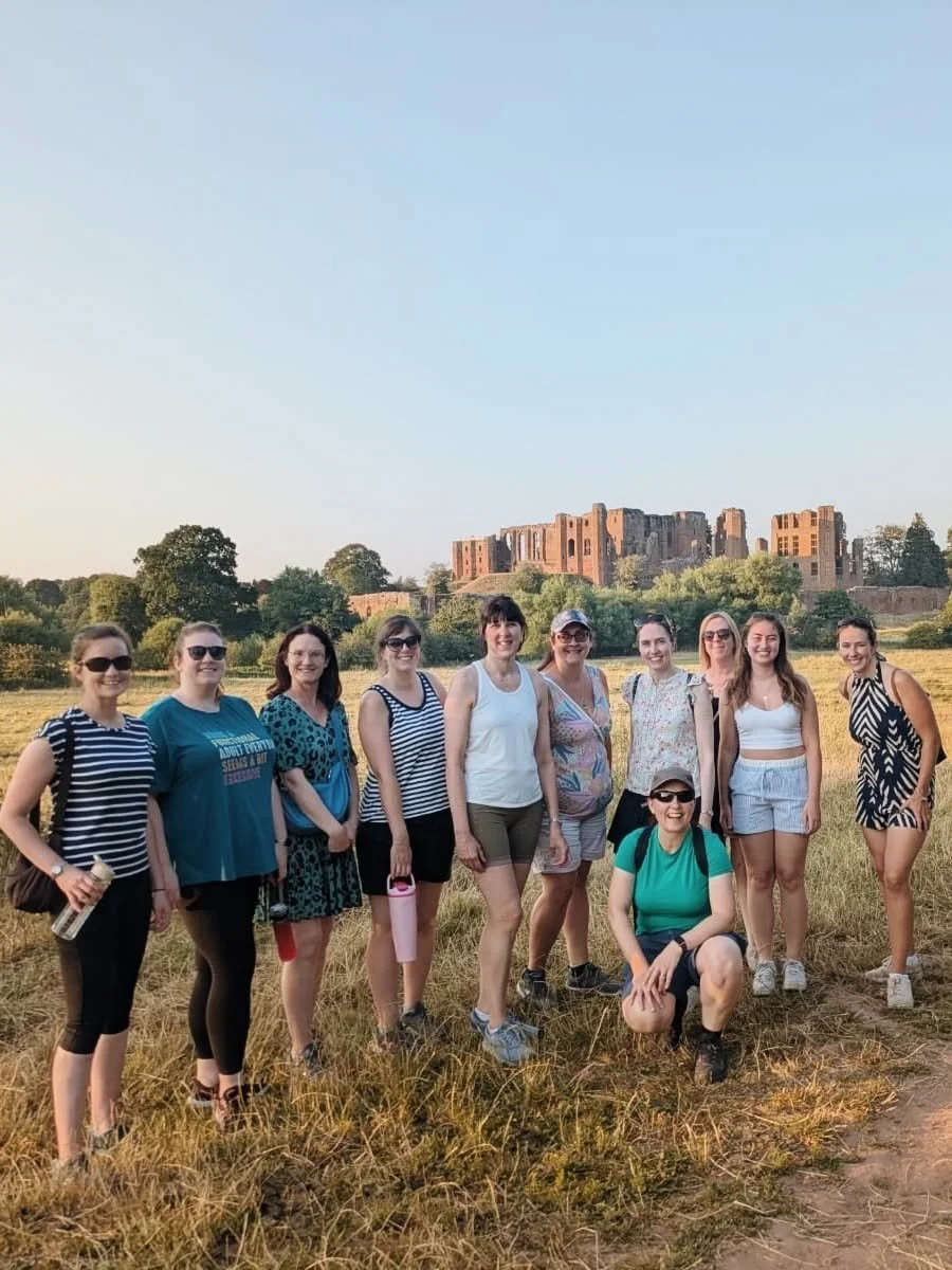 Group of ten women standing outdoors on a field with a historic castle in the background on a clear day.