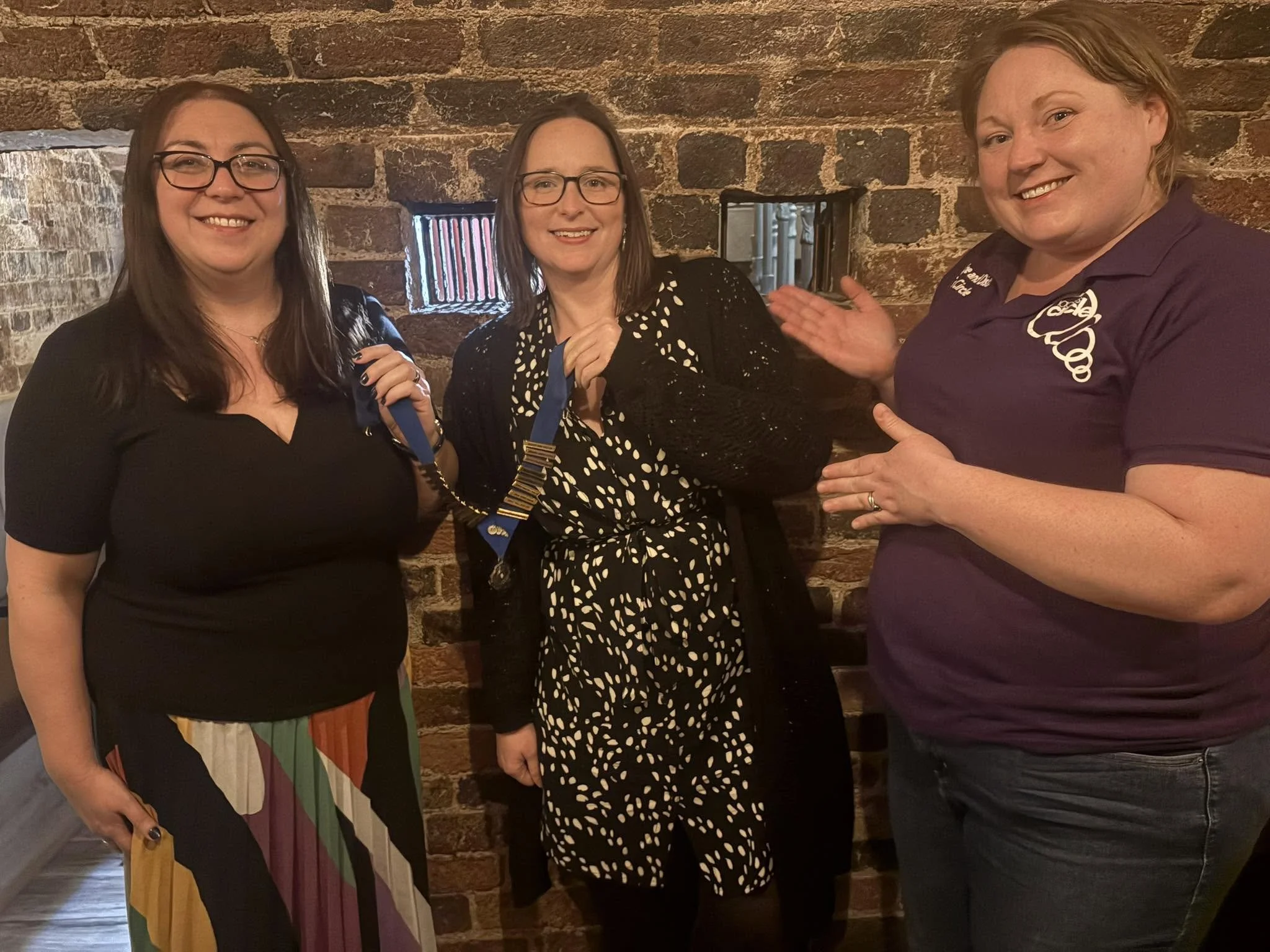Three women smiling and posing in front of a brick wall, with one woman holding a medal.
