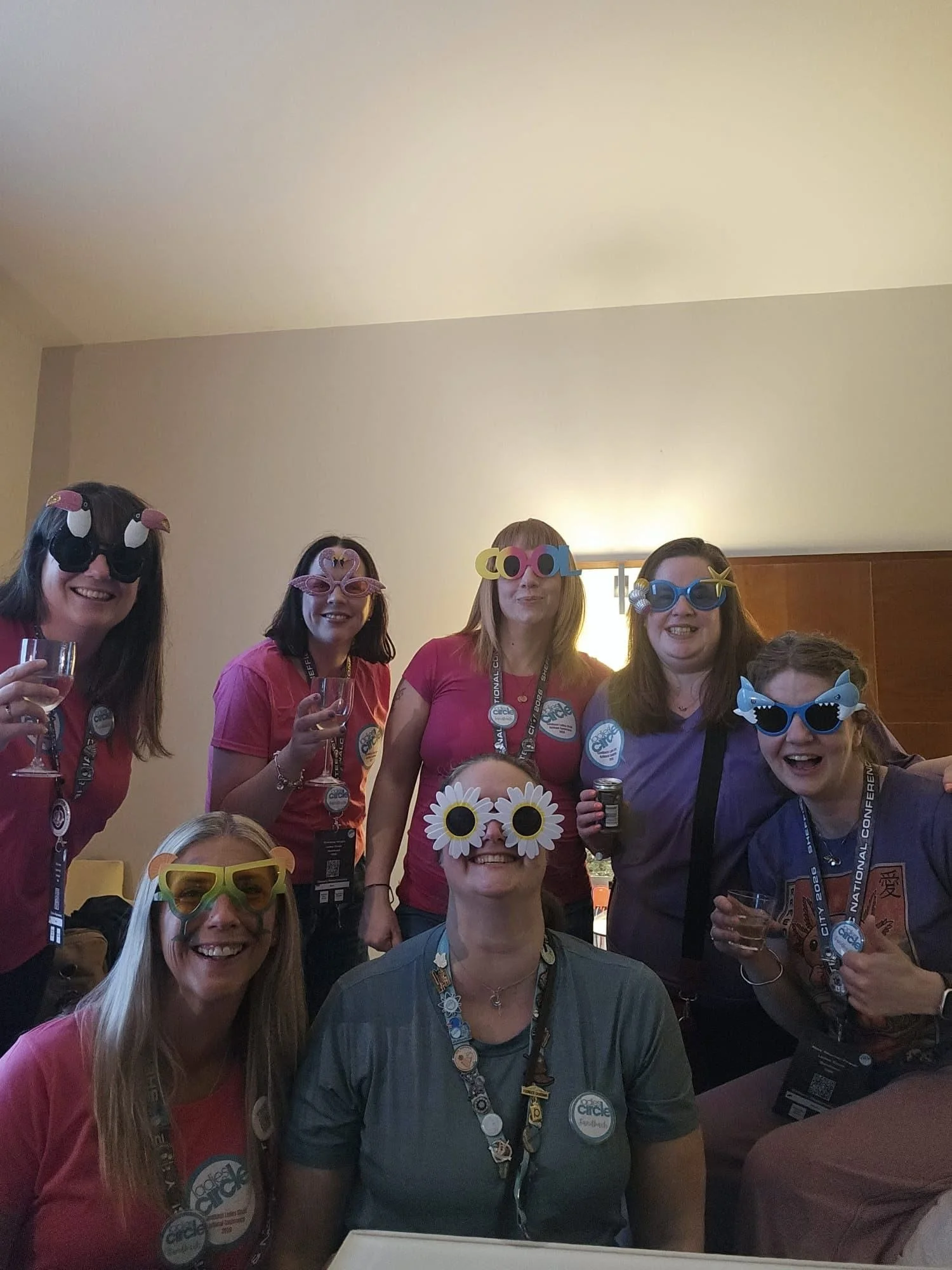 A group of people smiling, wearing colorful glasses and badges, at a conference or social gathering.