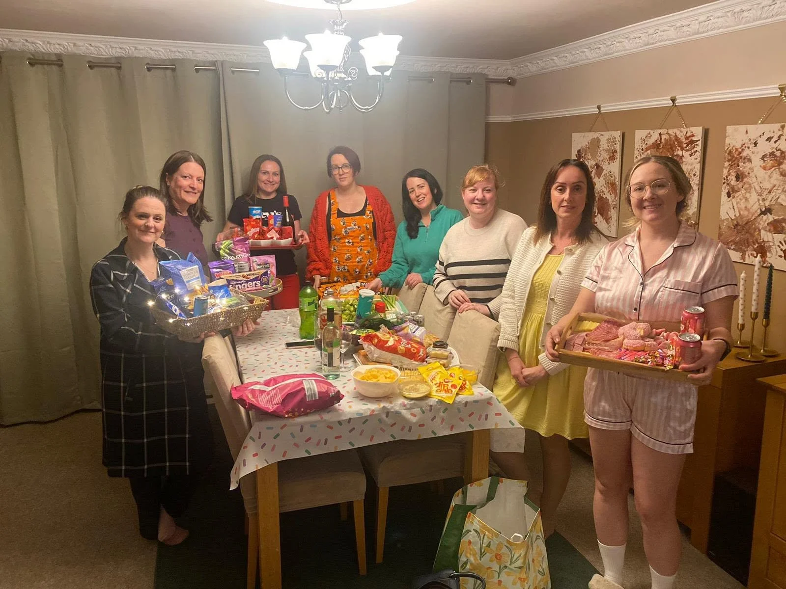 A group of nine women gathered around a dining table celebrating, with food and snacks on the table and walking around the table is a woman in pajamas holding a tray of meat and drinks.