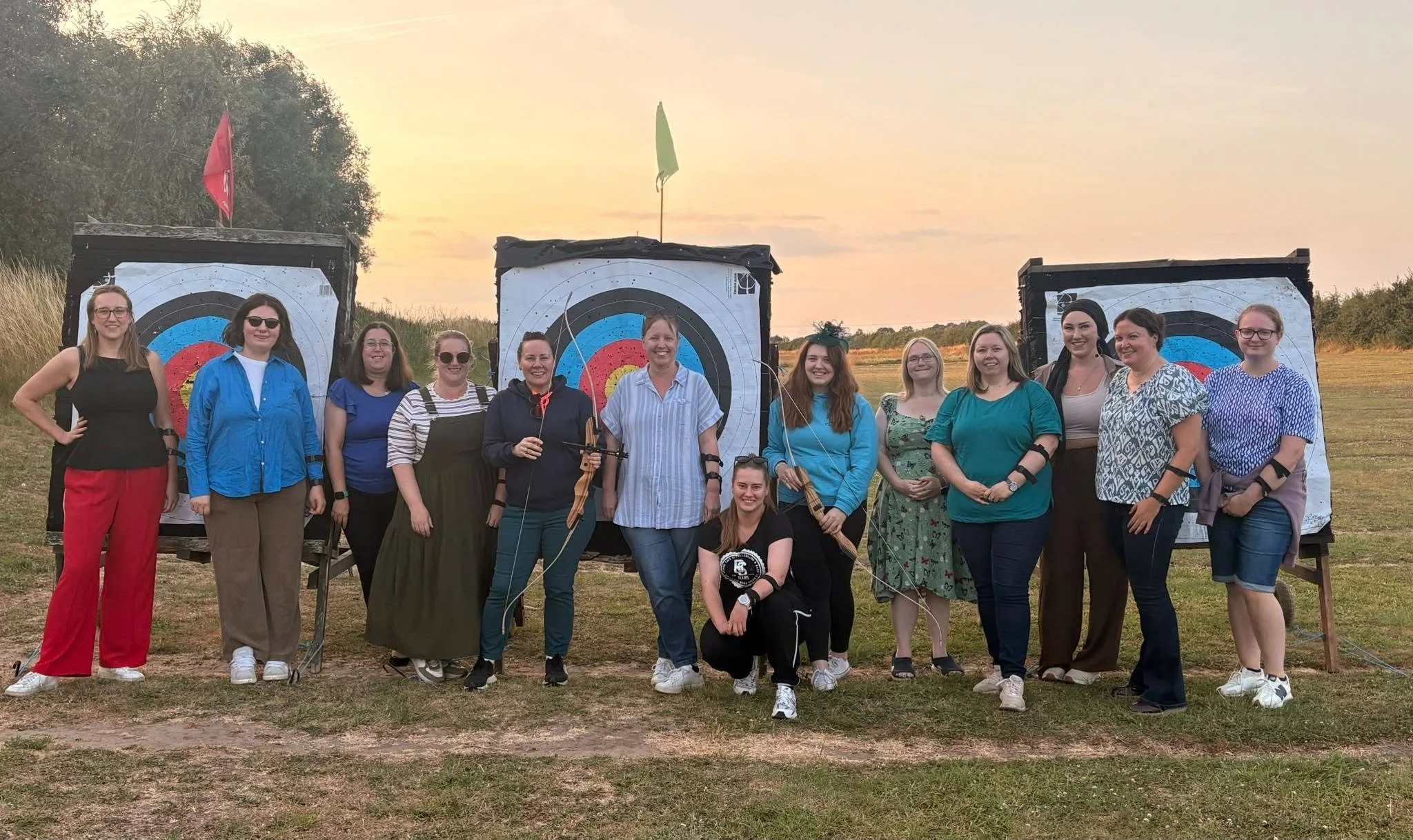 A group of fifteen women standing outdoors in front of three archery target boards during sunset, some holding archery bows and arrows, smiling at the camera.