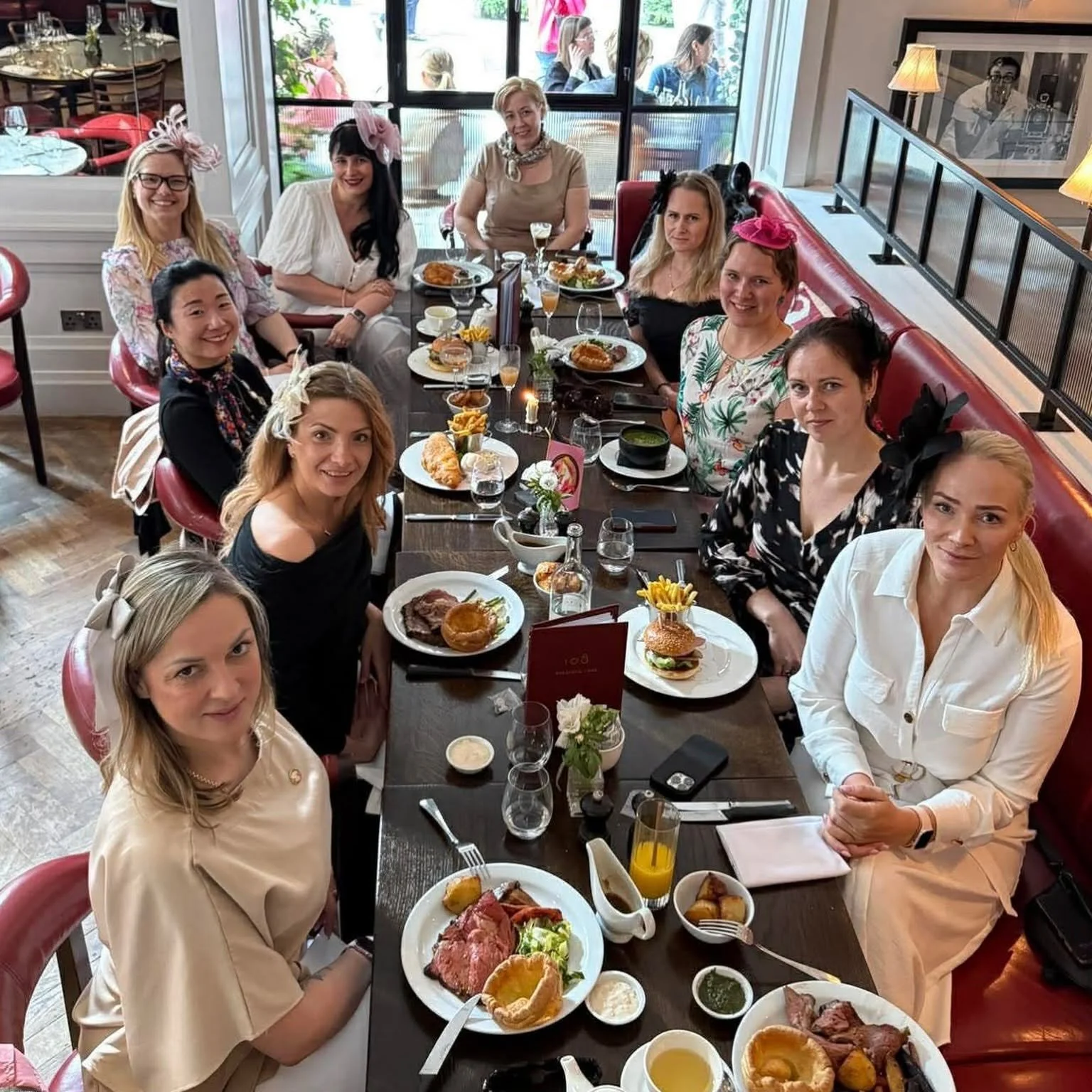 A group of ten women seated around a long dining table at a restaurant, enjoying a meal together. The table is set with plates of various foods, drinks, and small flower arrangements. Several women are wearing fancy headbands or hats, and some are smiling at the camera. The restaurant interior features large windows, red seating, and wall decor.