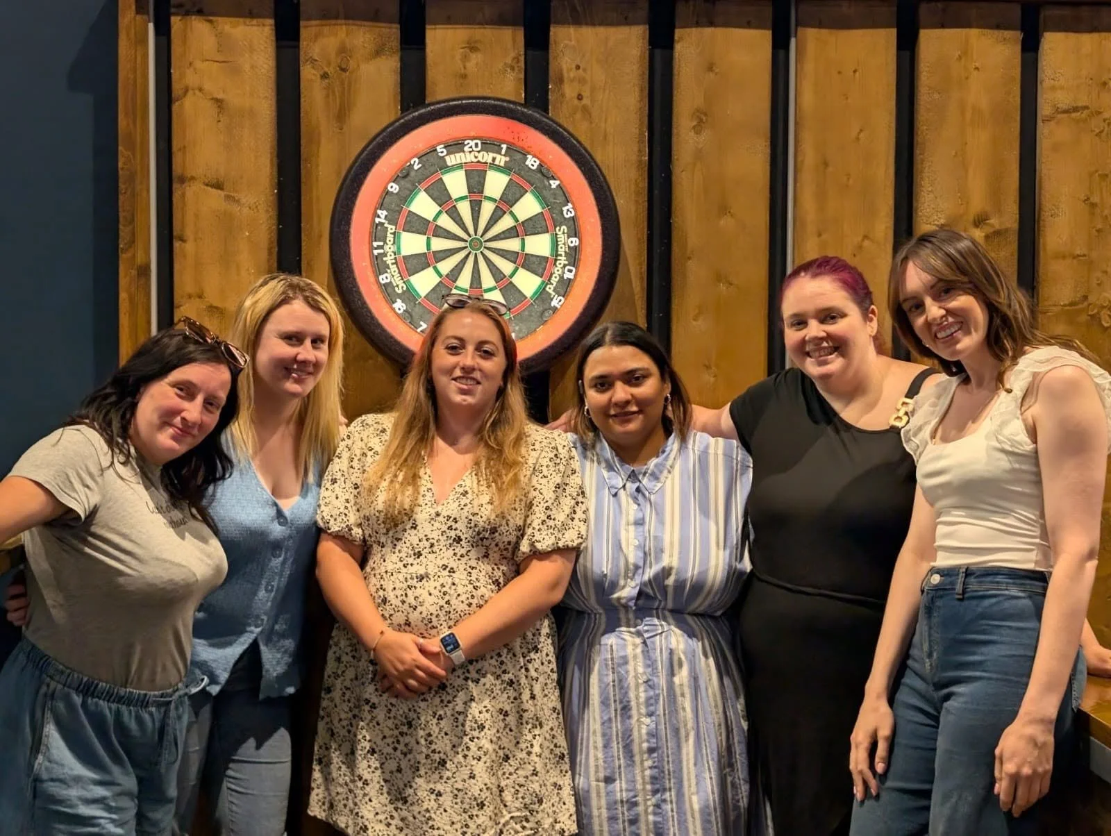 Six women standing in front of a dartboard, smiling for the camera, in a bar or pub setting.