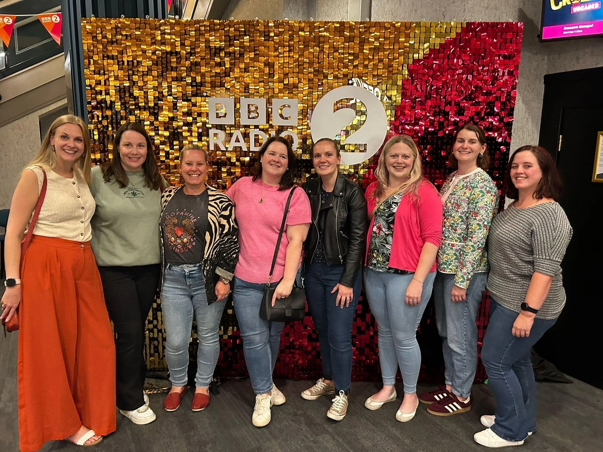 A group of eight women standing in front of a colorful, sequined backdrop with the BBC Radio 2 logo. They are smiling and dressed casually.