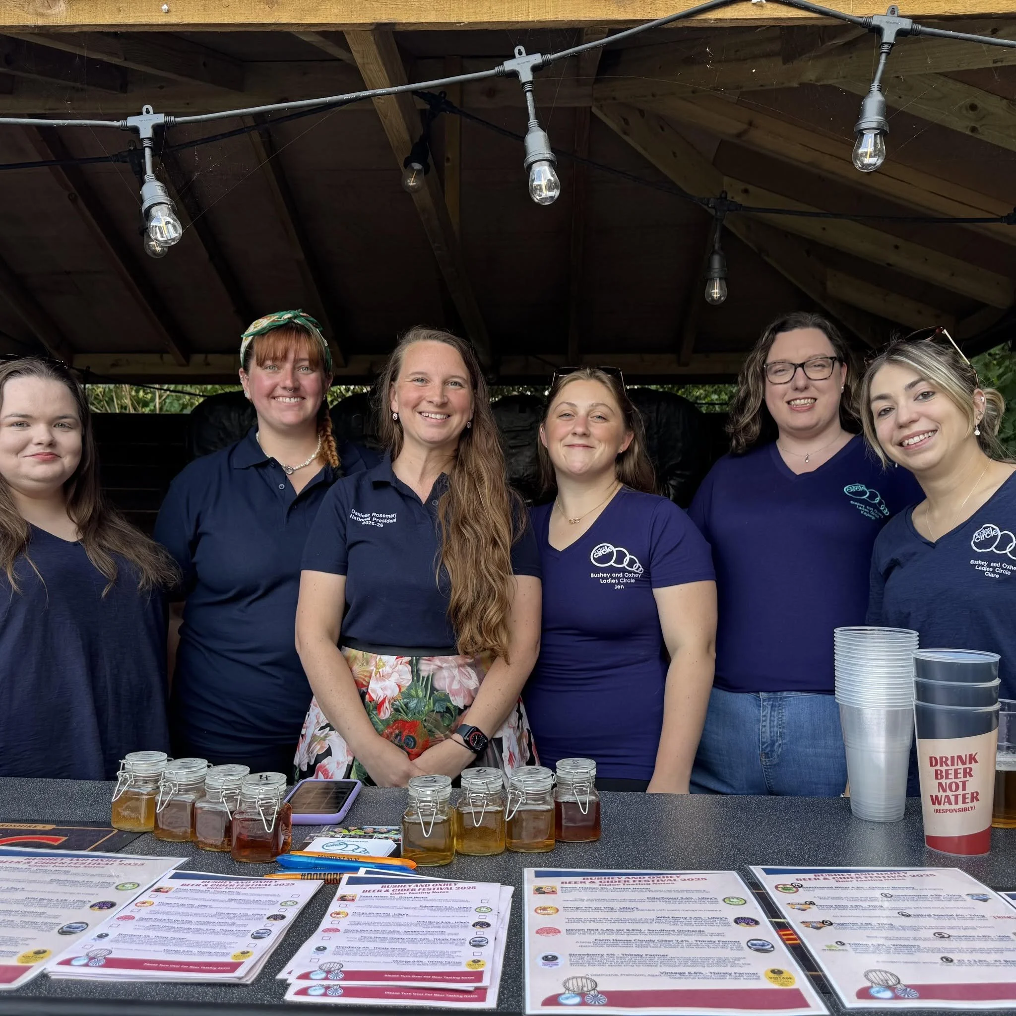 Group of women at an outdoor event under a wooden canopy, with various informational papers and jars on a table in front of them, and string lights hanging above.