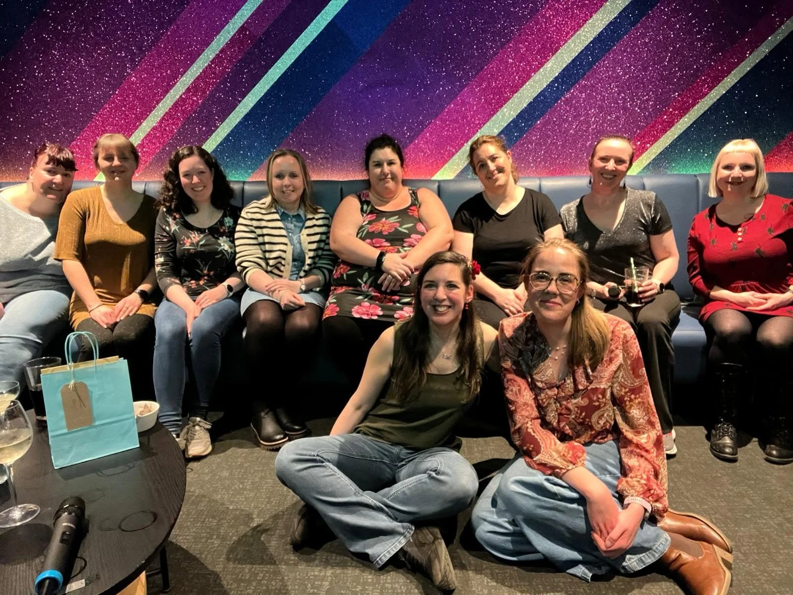 Group of eleven women sitting and kneeling in front of a vibrant, colorful, starry wall with diagonal stripes, celebrating together at a party.