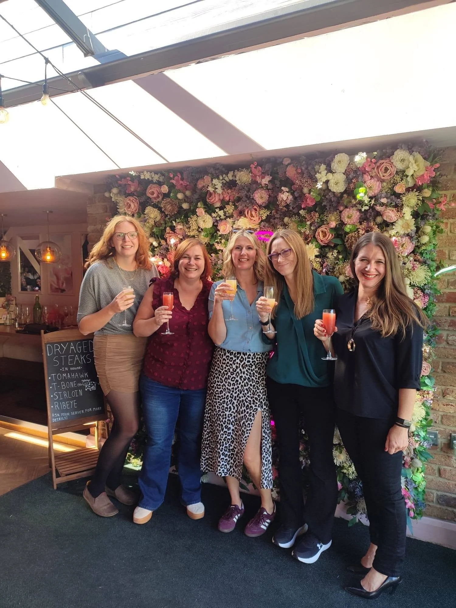 Five women standing together, smiling and holding drinks, in front of a floral backdrop in an indoor event space.