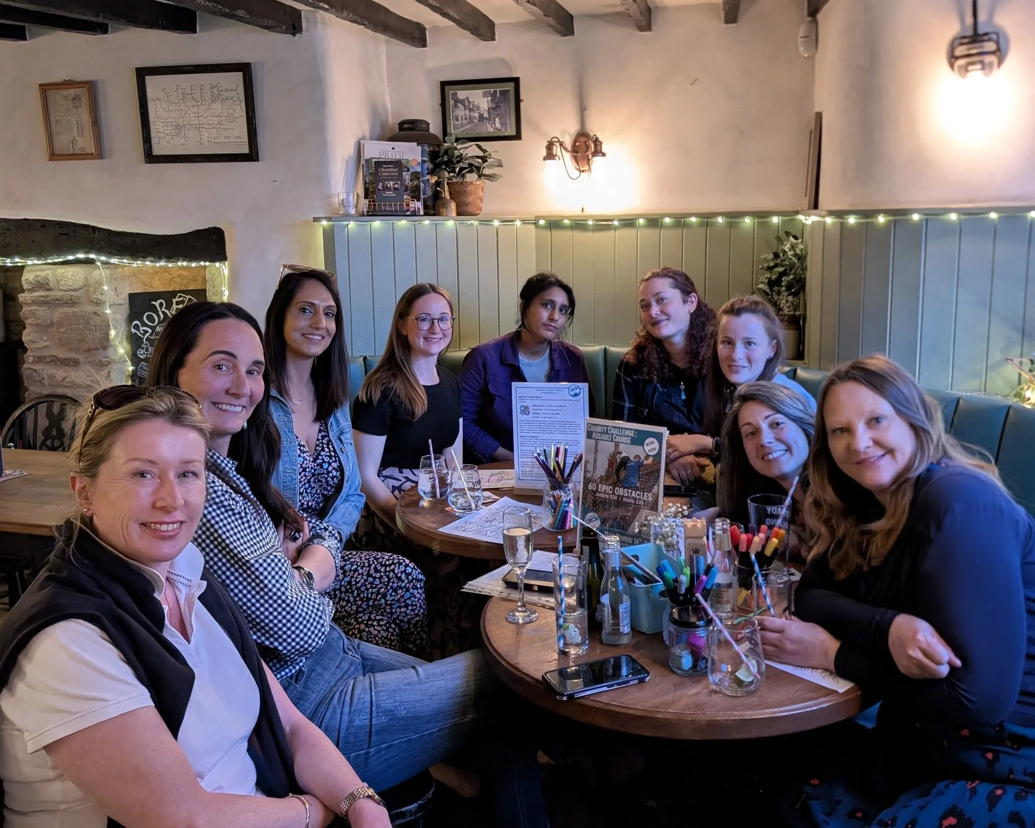 A group of women sitting around a table in a cozy restaurant or cafe, smiling at the camera. The table has drinks, pens, and a sign, with a decorated wall and pictures in the background.