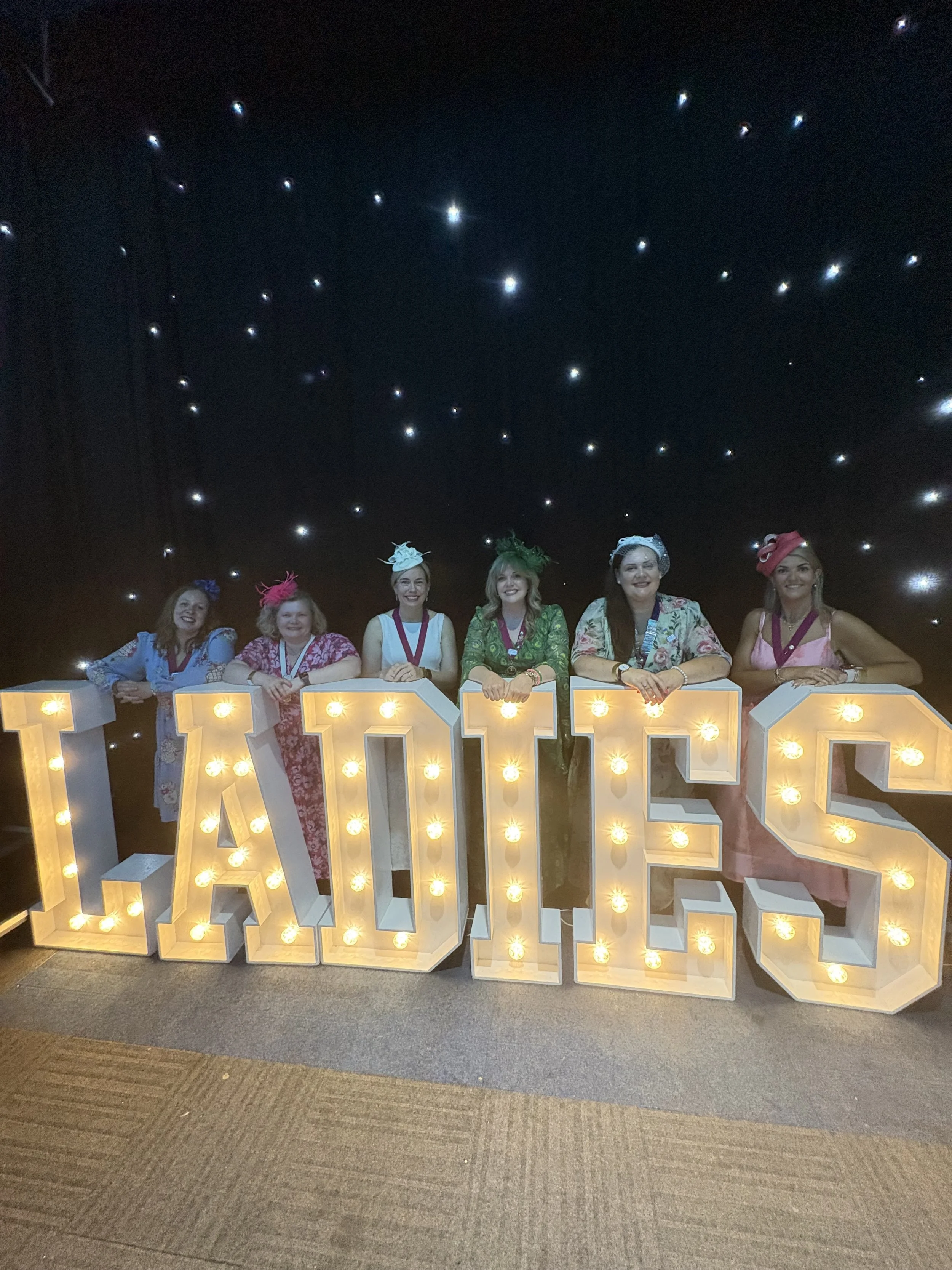 Group of six women wearing colorful hats standing behind illuminated letters spelling LOVE.