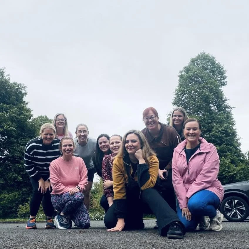 A group of ten women smiling outdoors on a cloudy day, with trees and a car in the background.
