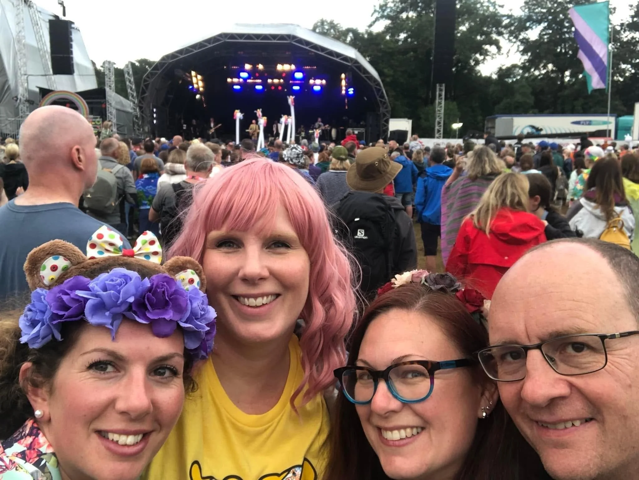 Group of people at an outdoor concert, woman with pink hair smiling in the foreground, surrounded by others, some wearing glasses and colorful outfits.