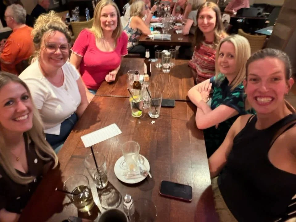 Group of women smiling and sitting around a wooden table at a restaurant or bar, with drinks and glassware on the table.
