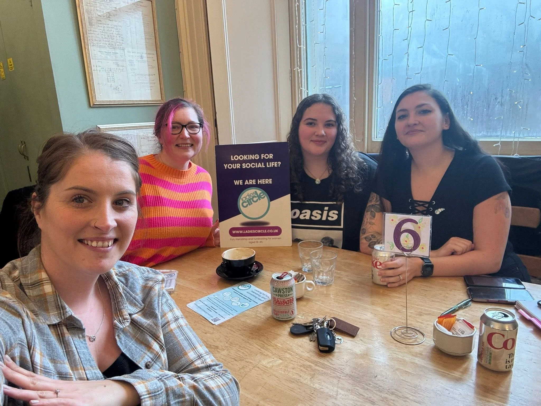 A group of four women sitting at a wooden table in a room with light green walls and a large window. They are smiling and looking at the camera. On the table are various items including a cup of coffee, a set of keys, papers, and a sign that says "Looking for your social life? We are here," with a website URL www.ladiescircle.co.uk.