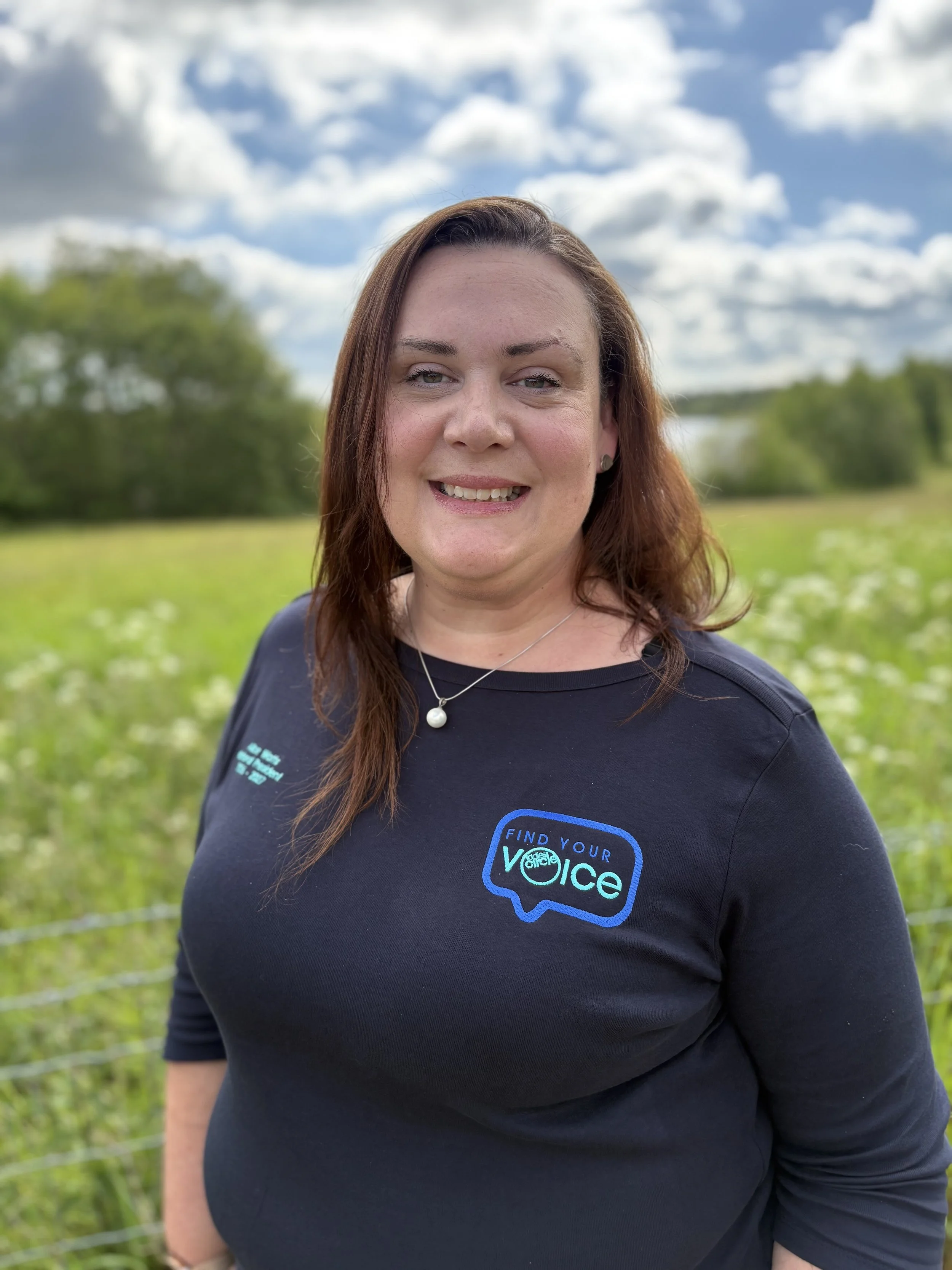 A woman with long brown hair smiling outdoors, wearing a navy blue shirt with a logo that says "Find Your Voice" and a pearl necklace, standing in a green field with trees and a blue sky with clouds in the background.