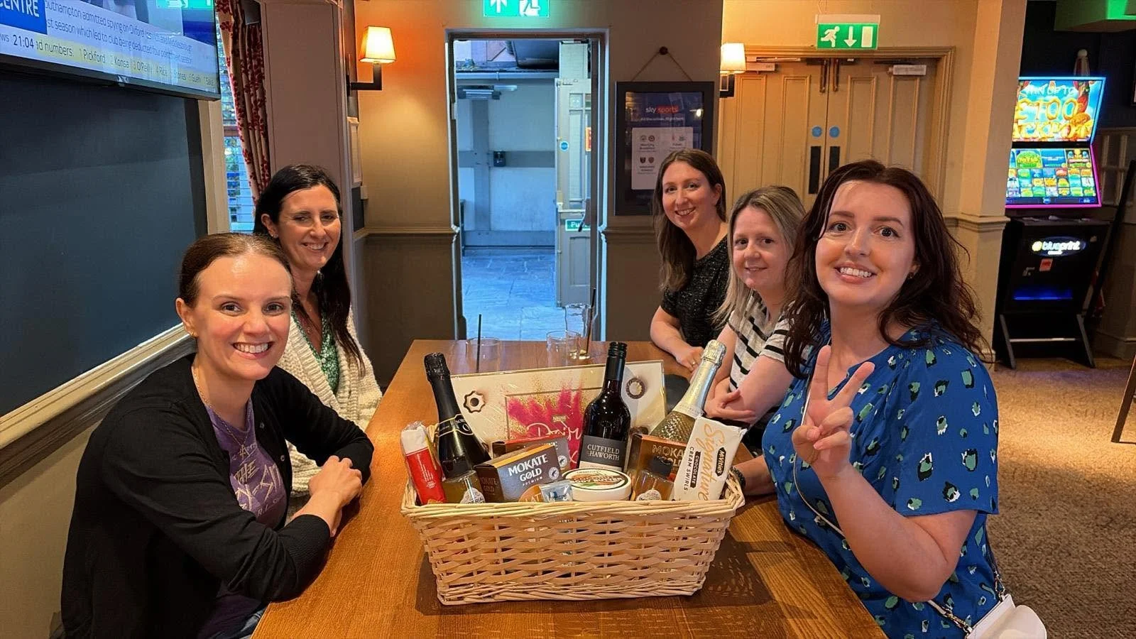 Five women sitting around a table with a gift basket in the center, smiling and celebrating in a bar or pub setting.