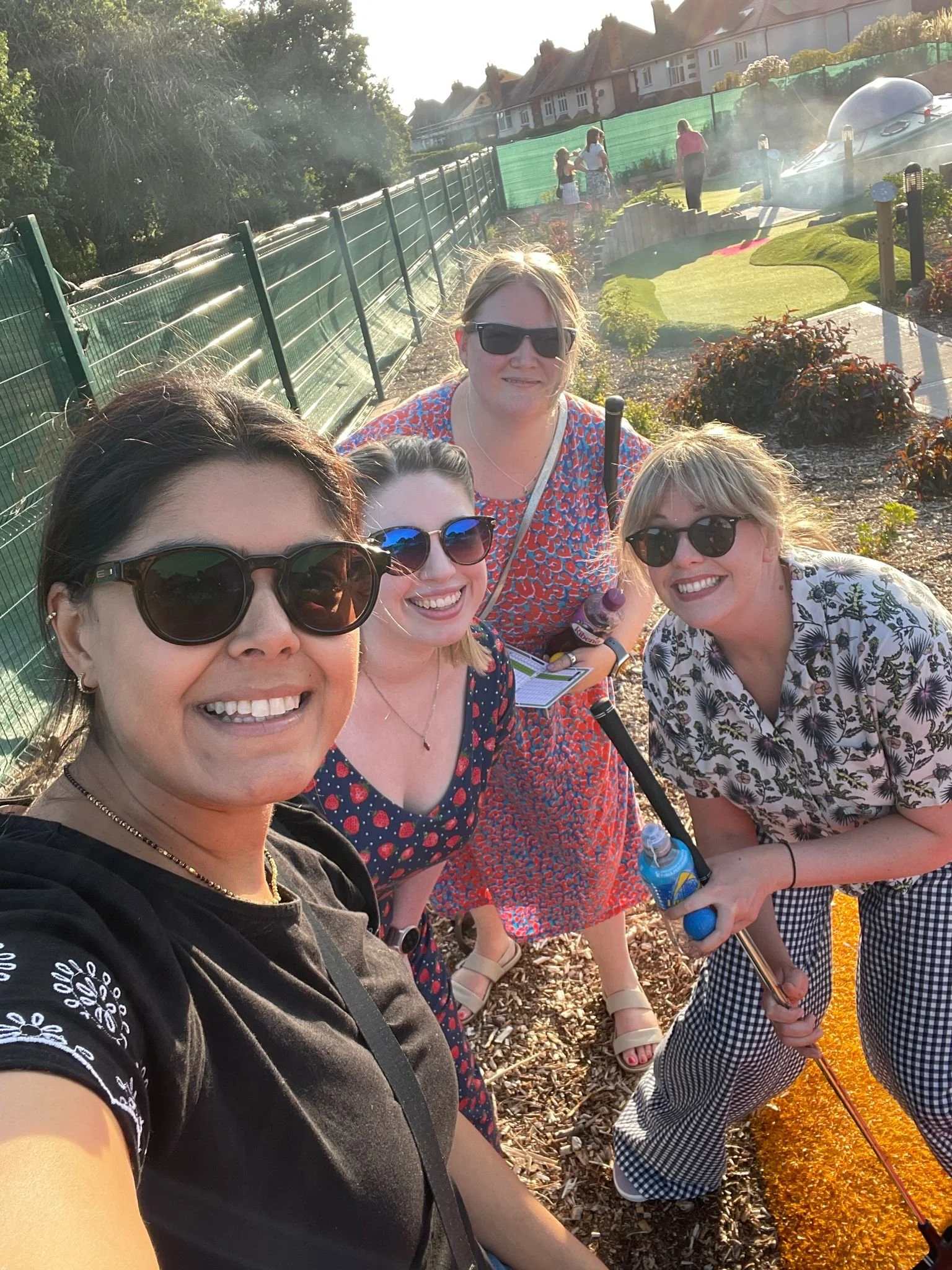 Group of four women taking a selfie outdoors in a garden area during late afternoon, with a decorative winding garden bed and a fence in the background.