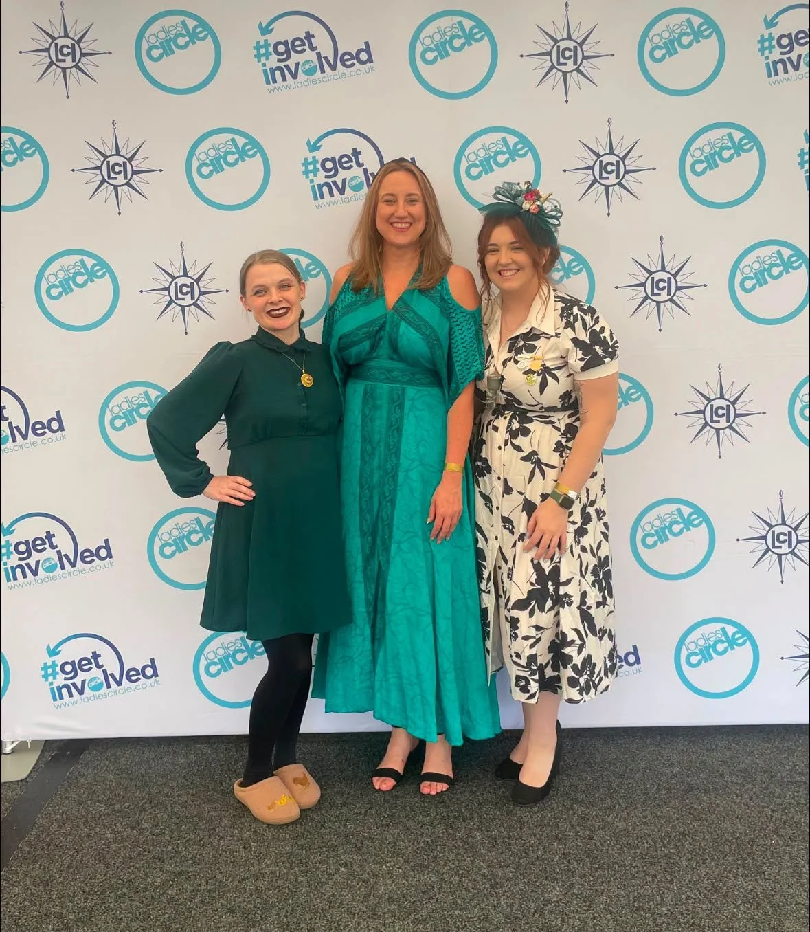Three women smiling and posing for a photo in front of a backdrop with logos and hashtags, including #getinvolved and Ladies Circle.