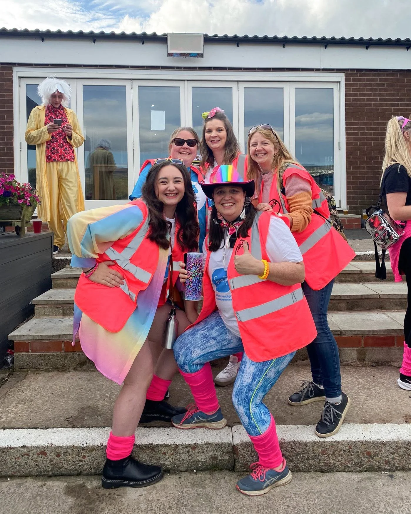 Group of five women wearing colorful clothing and reflective safety vests, smiling and posing on outdoor steps in front of a building with glass doors. One woman in the background is dressed in a yellow outfit, wig, and holding a phone.