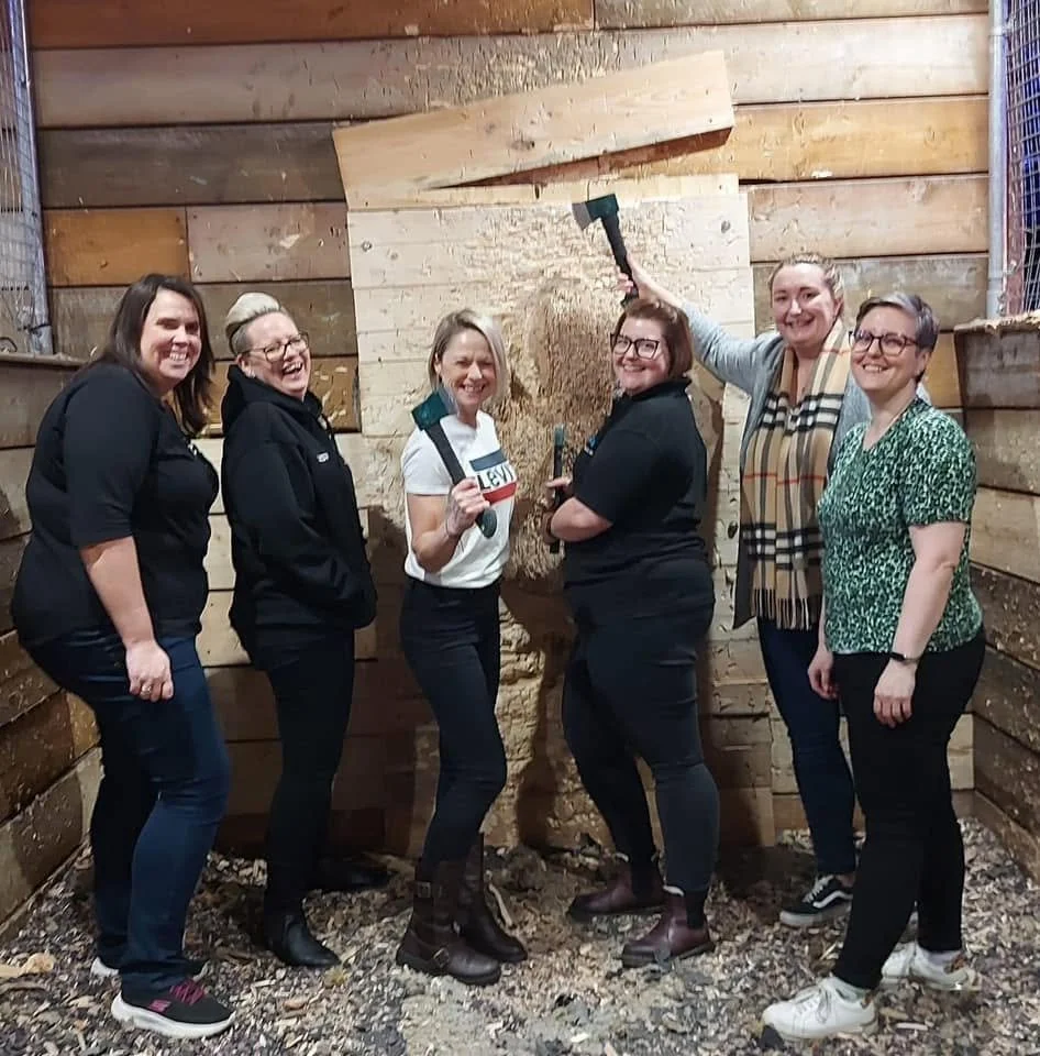 Six women standing inside a wooden shed, posing together and smiling. One woman is holding a sledgehammer raised above her head, and another woman is holding a hammer. They appear to be celebrating or completing a woodworking or renovation project.