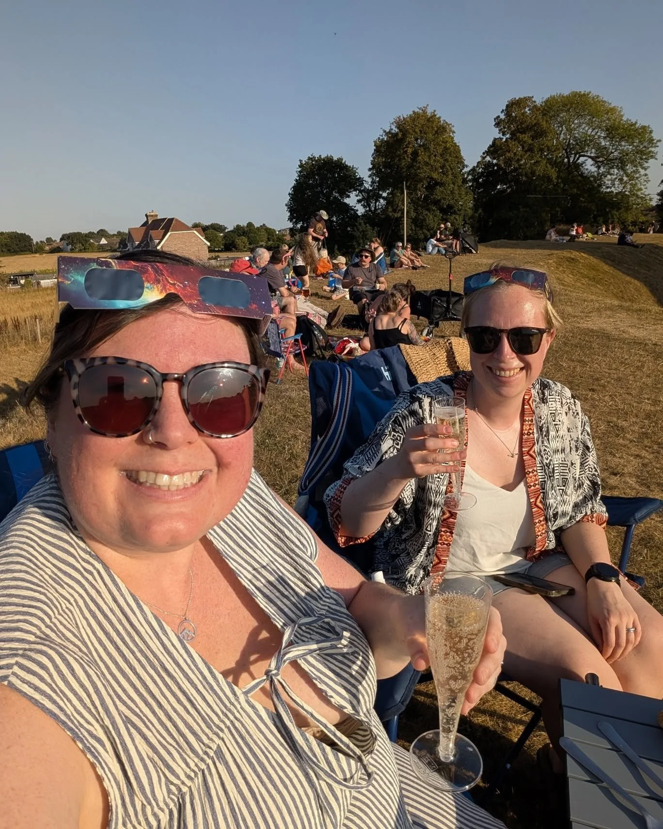 Two women seated outdoors smiling at the camera, one holding a glass of champagne. In the background, people are sitting on the grass and enjoying the sunny weather in what appears to be a park or open field.