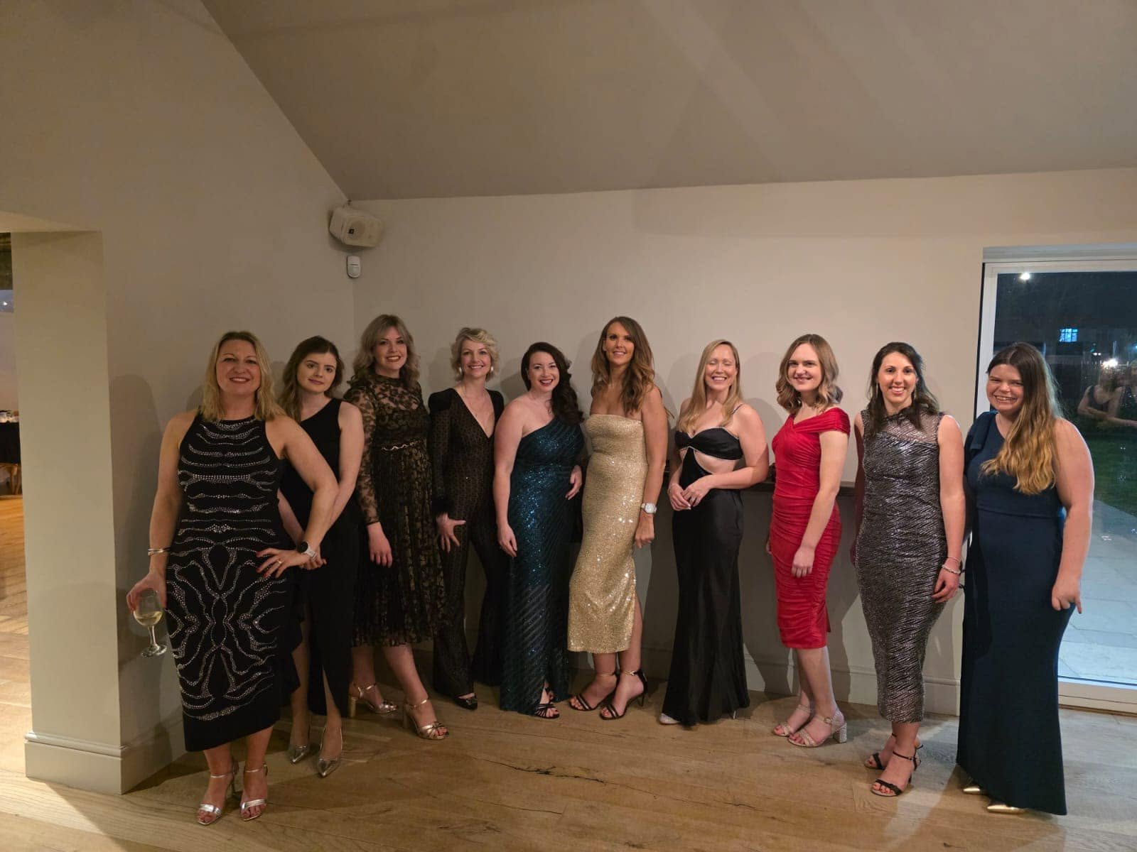 A group of eleven women dressed in formal evening gowns, standing together indoors and smiling for the camera. One woman on the far left is holding a glass of white wine.