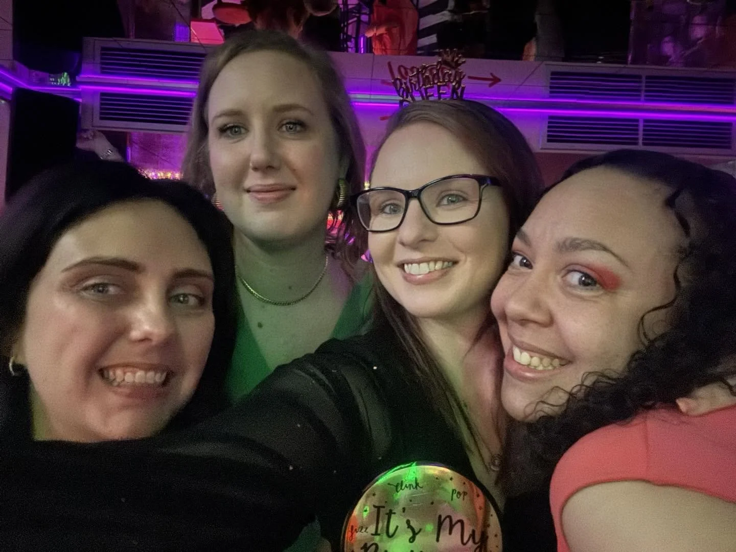 Four women taking a group selfie at a party with colorful lighting and a sign in the background that says "happy birthday."