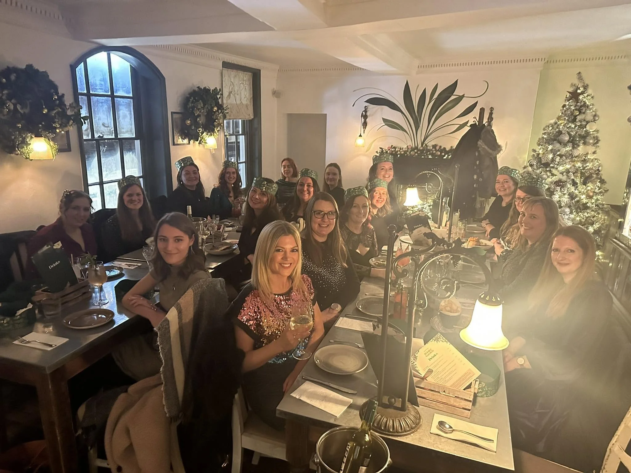 Group of women sitting at a restaurant table, dressed festively, celebrating during the holiday season. There are Christmas trees and holiday decorations in the background.