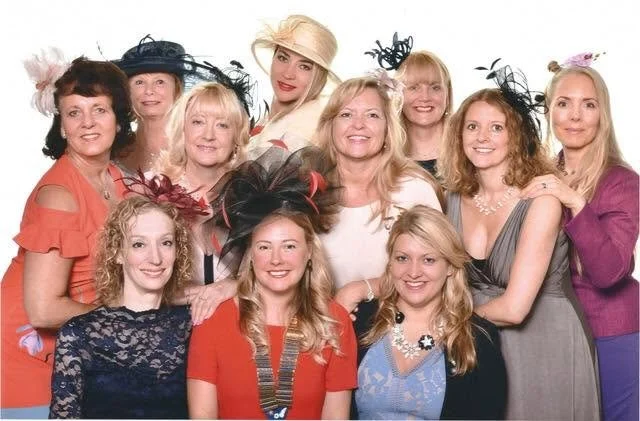Group of twelve women dressed in colorful and fancy outfits, wearing various hats and accessories, smiling together for a photo.