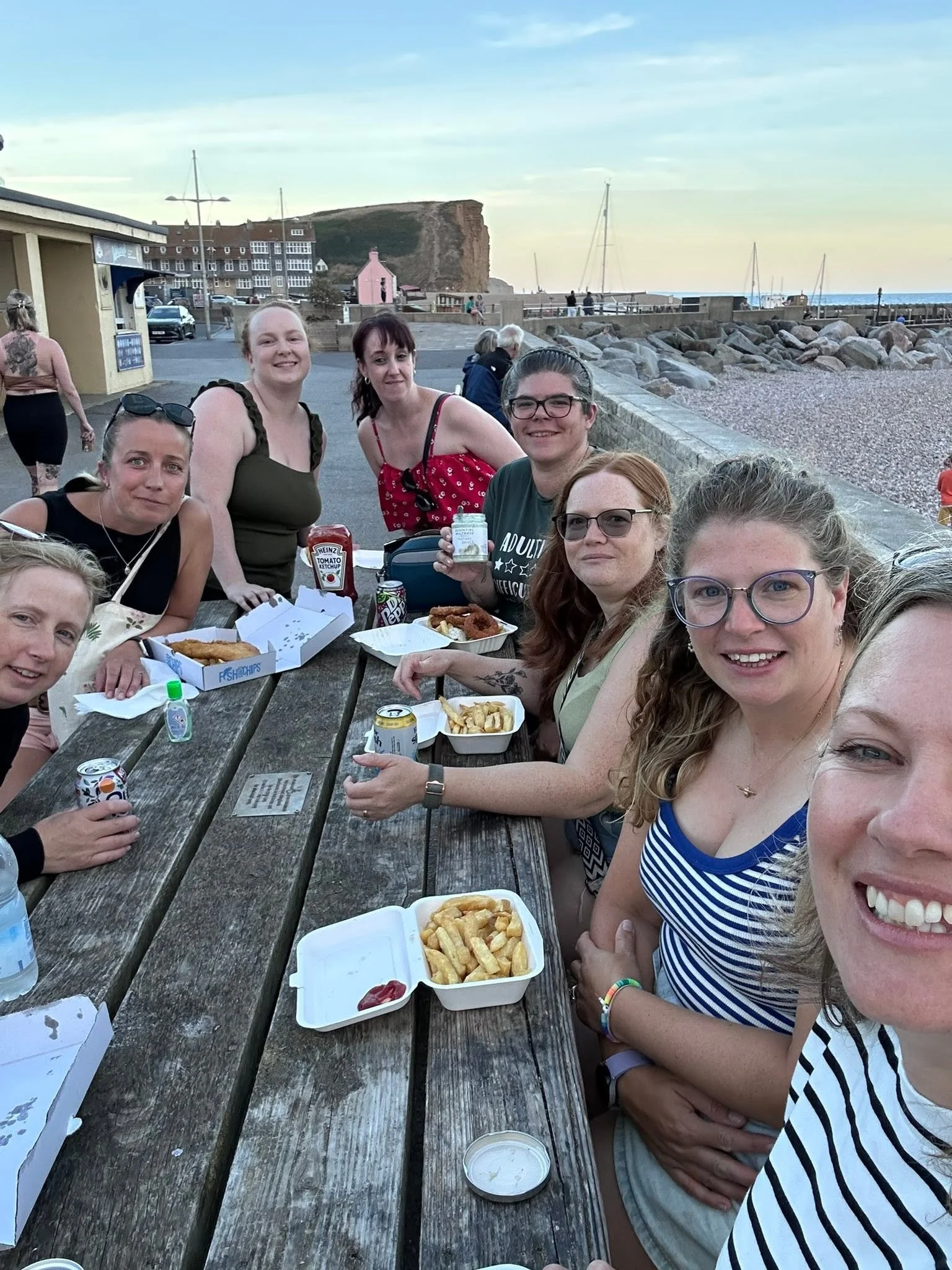 A group of people sitting at a wooden outdoor table near a beach, enjoying food and drinks, with boats and a rocky shoreline in the background.