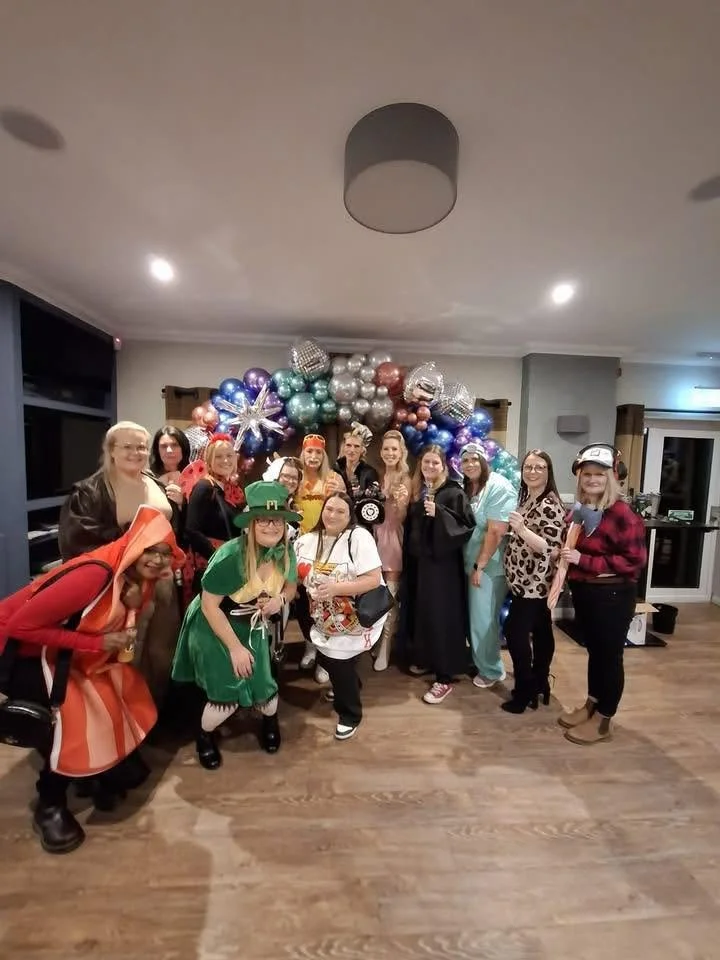 Group of people dressed in costumes standing in front of a balloon arrangement with star-shaped balloons, at a party or celebration.